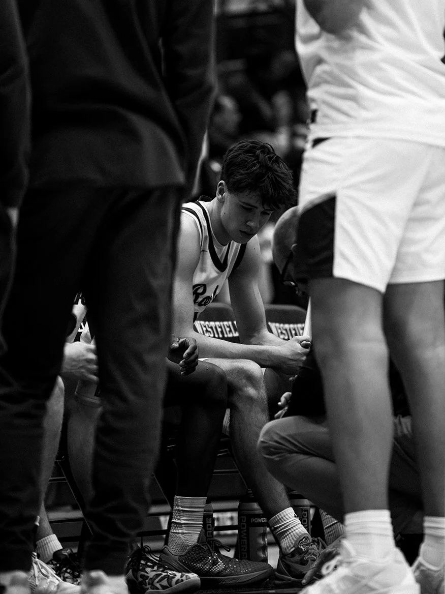 A basketball player sitting on the bench during a game, surrounded by coaches and teammates, appearing focused and serious.