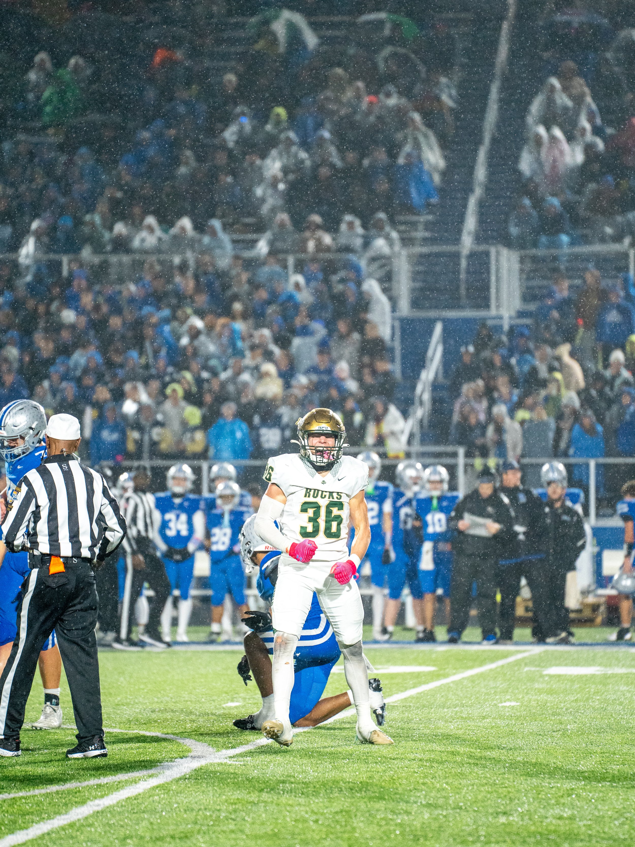 A football game in rain, with a player in a white and gold uniform standing on the field, surrounded by players and officials, with a crowd in the stands in the background.