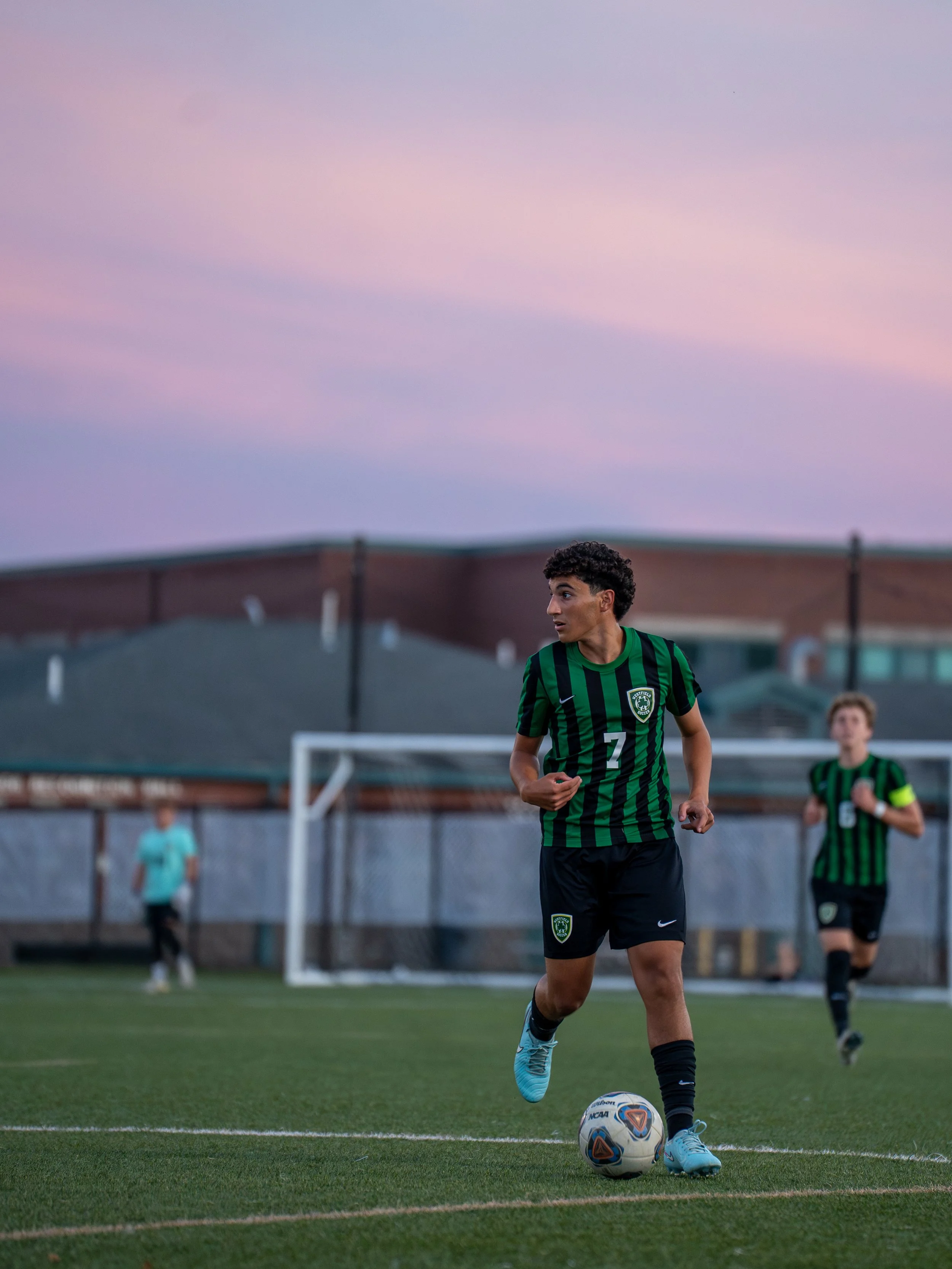 A young male soccer player wearing a green and black striped jersey with the number 7 is on the field with a soccer ball at his feet. In the background, there are other players, a goalpost, and a fence, with a twilight sky overhead.