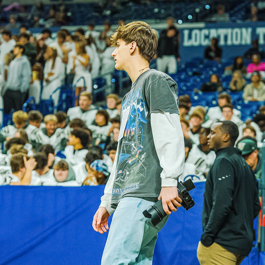 A young man with brown, wavy hair holding a camera with a telephoto lens at a sports event, standing near a blue barrier, with a crowd of spectators and players in hockey jerseys in the background.