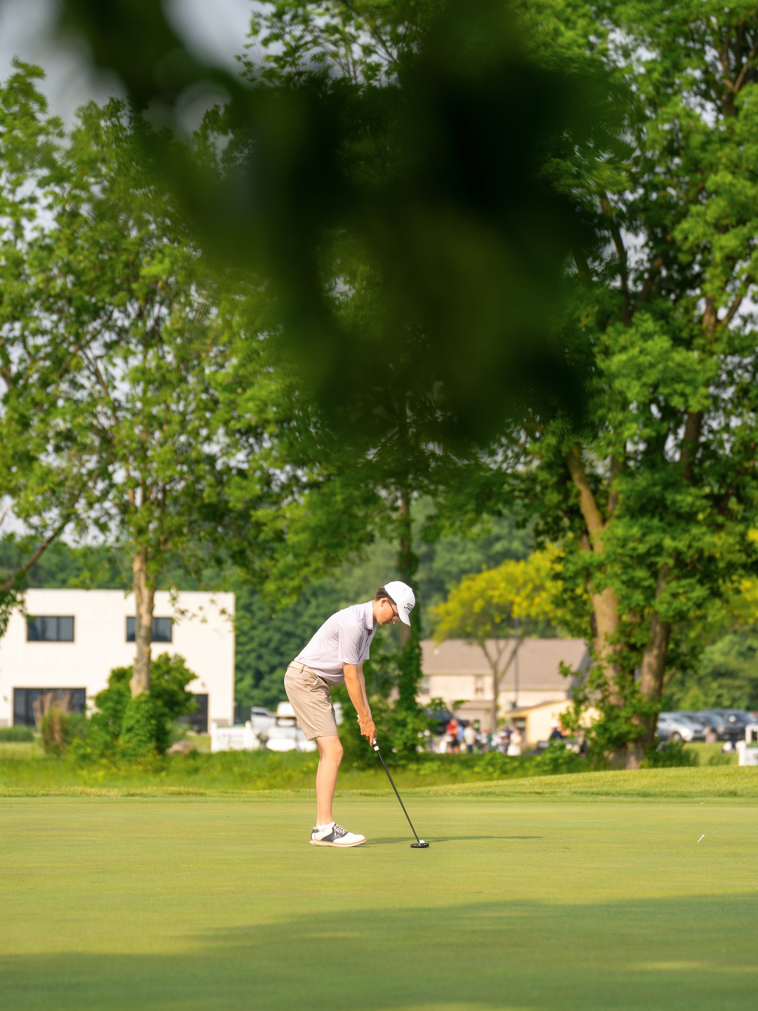 A person in a white cap and pink shirt playing golf on a green course, with trees and buildings in the background, seen through a blurred branch.