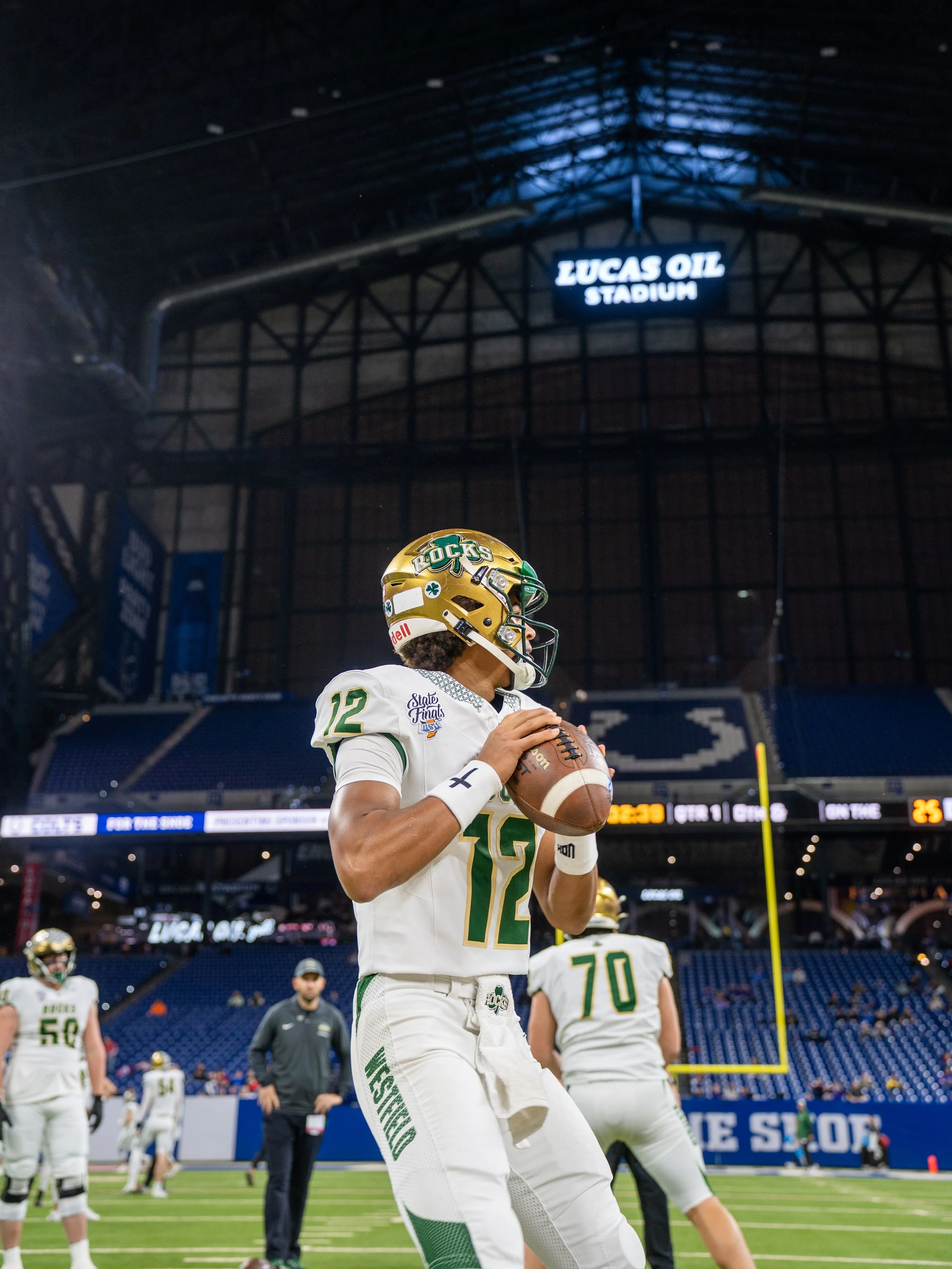 A football player wearing a white and green uniform with the number 12, holding a football and preparing to throw, on a field at Lucas Oil Stadium.