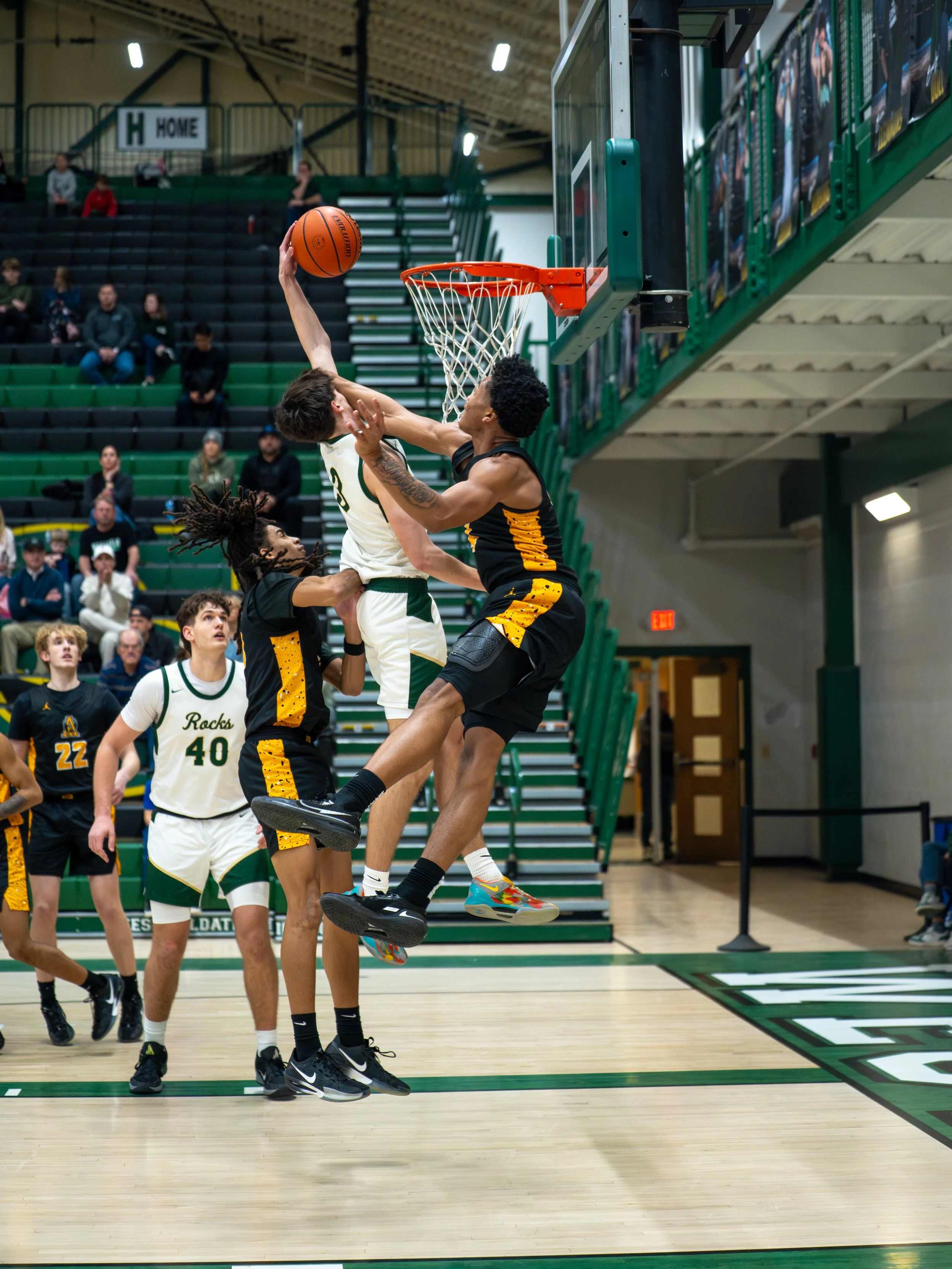 Two basketball players are jumping near the hoop, one attempting a shot while the other tries to block. Several other players and spectators watch from the background in an indoor basketball court.