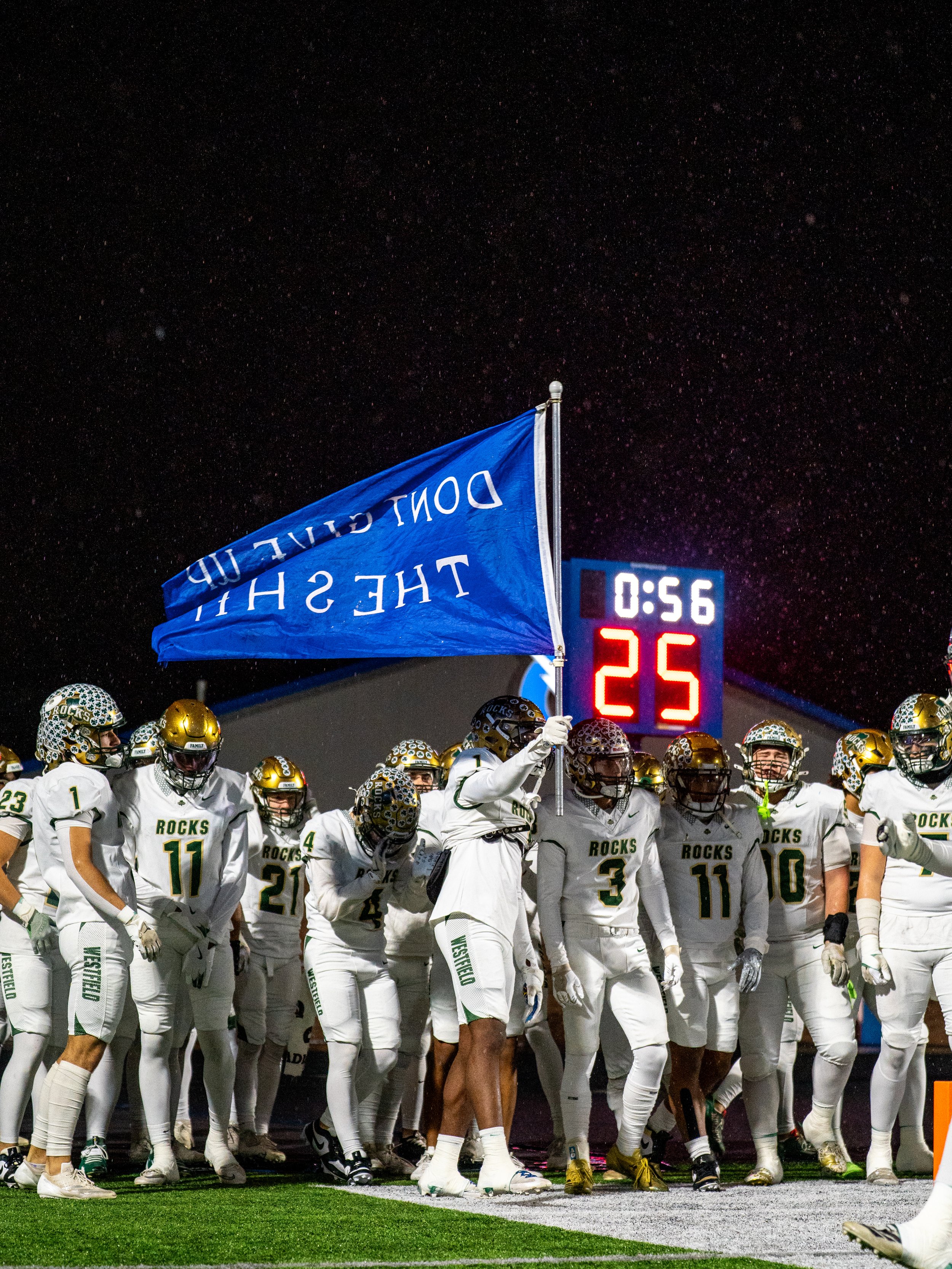Football team called the Rocks standing on the field, holding a blue flag, and preparing for the game in the rain at night, with a scoreboard showing 0:56 time and 25 points.