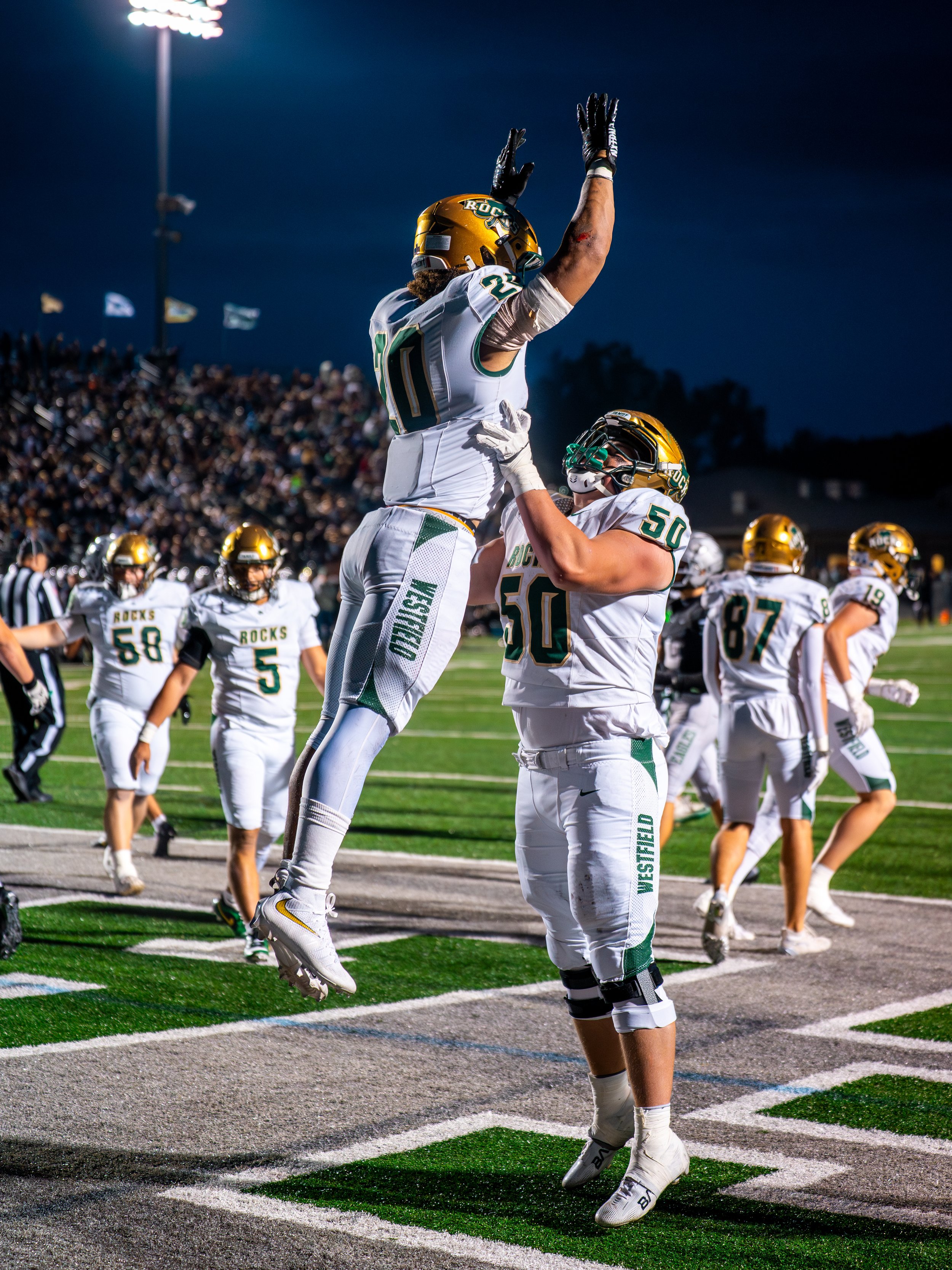 Football players in white and green uniforms celebrating on the field under stadium lights during a game, with one player jumping after scoring, and fans in the stands in the background.