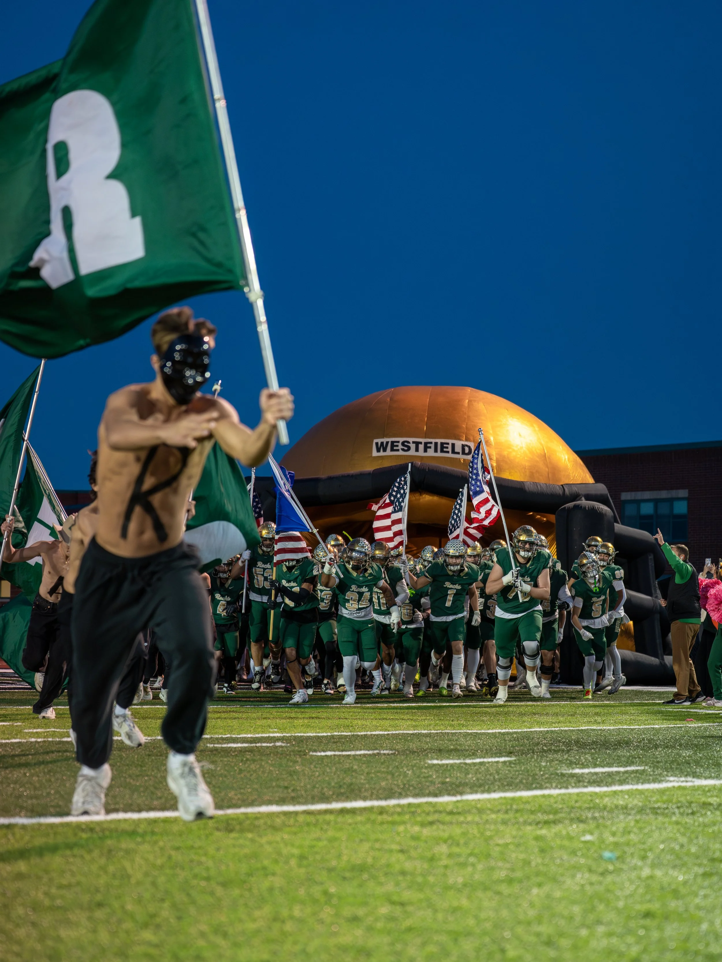 High school football team running onto the field through a tunnel, holding flags and a large inflatable helmet with the label 'Westfield', on a football field at dusk.