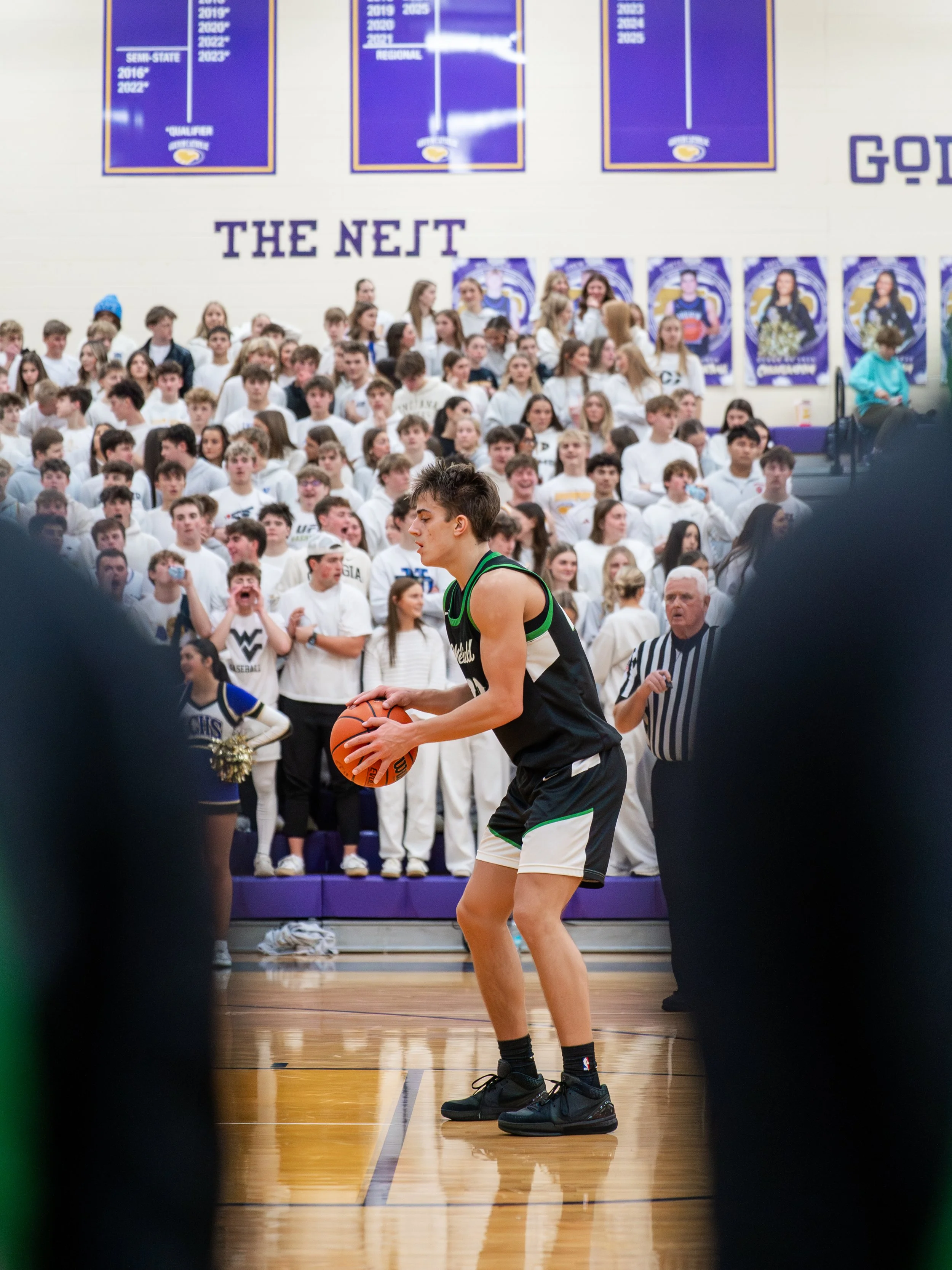 A young basketball player wearing a black and green uniform preparing to shoot a free throw on a gymnasium court, with a crowd of spectators and classmates in white shirts watching from the bleachers.