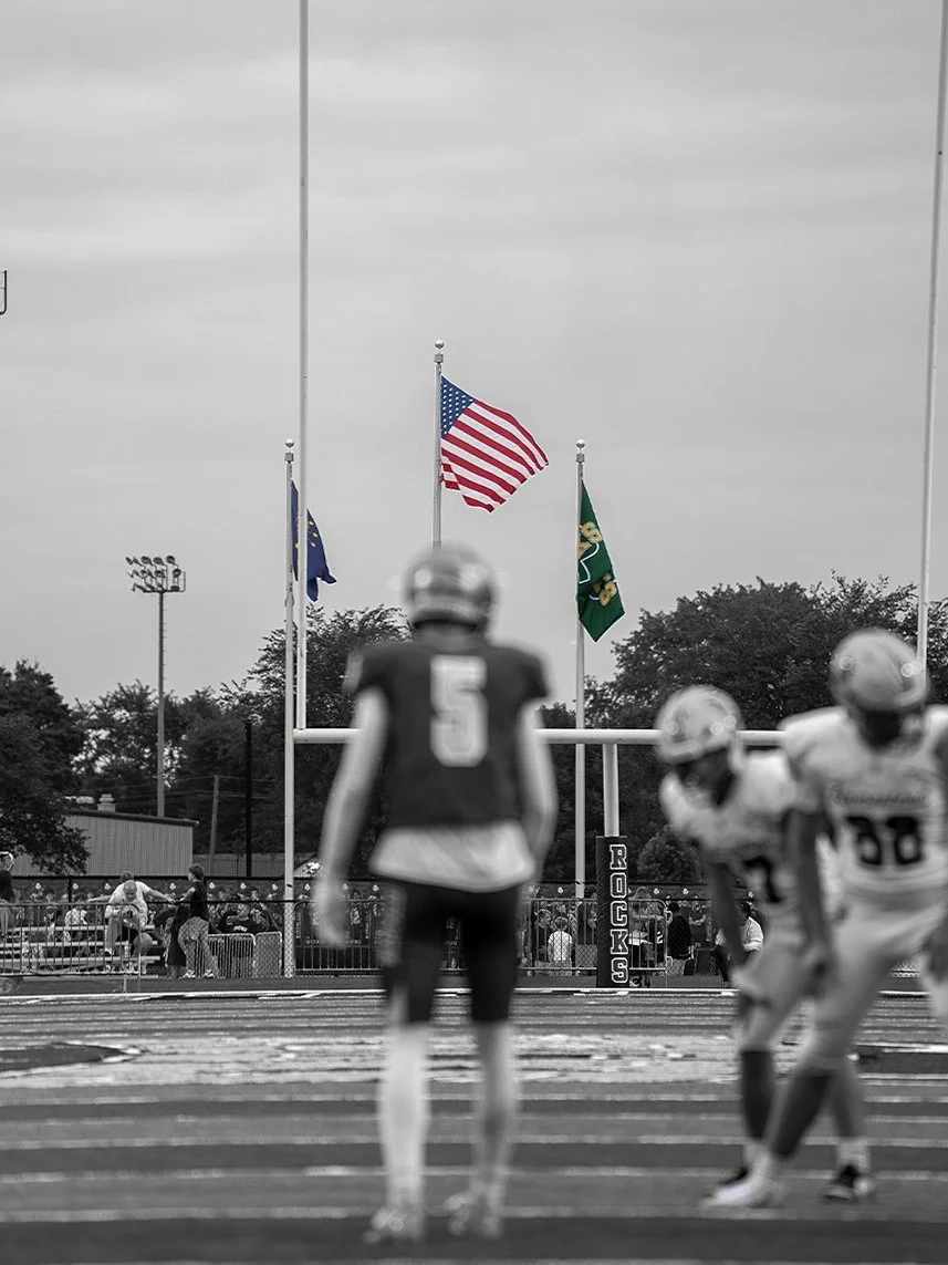 Football game on field with American flag, flags, and players in helmets, with spectators in the background.