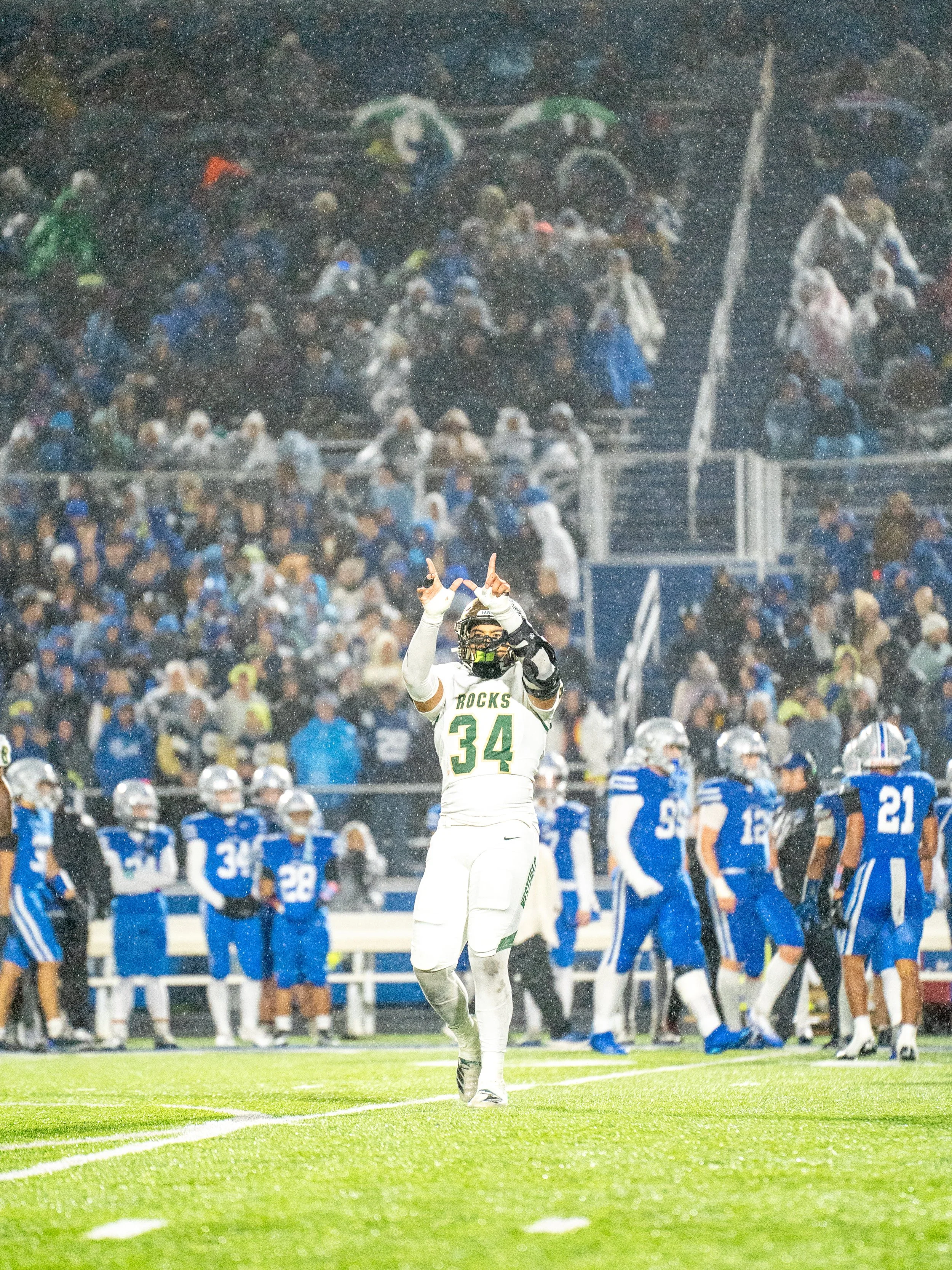 A football player in a white and green uniform celebrating on the field during a game with rain falling, and a crowd of spectators in the stands.