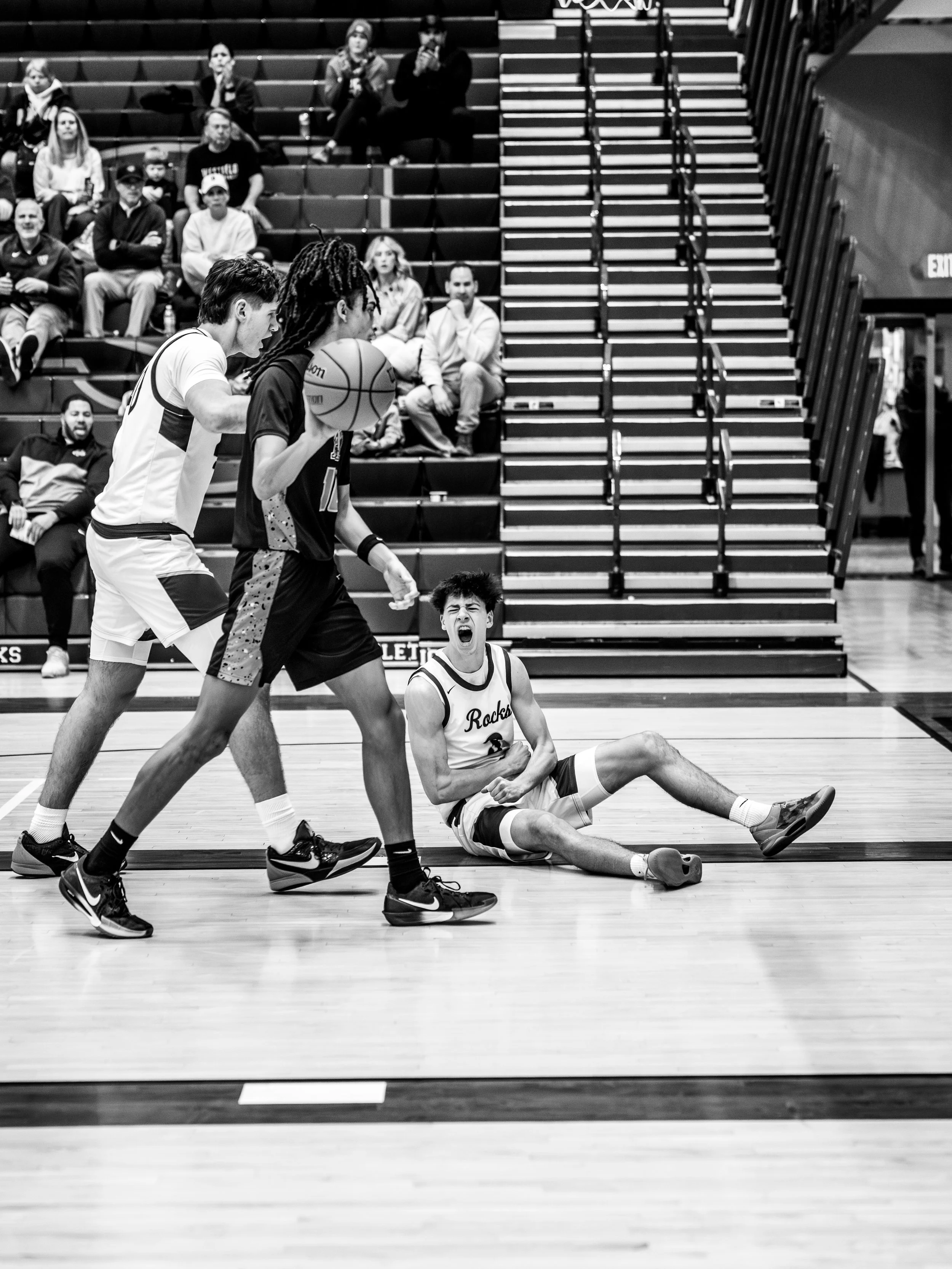 A basketball game where one player is lying on the floor, clutching his stomach and yelling, while two other players are standing nearby, one holding the basketball. Spectators are seated in the background.