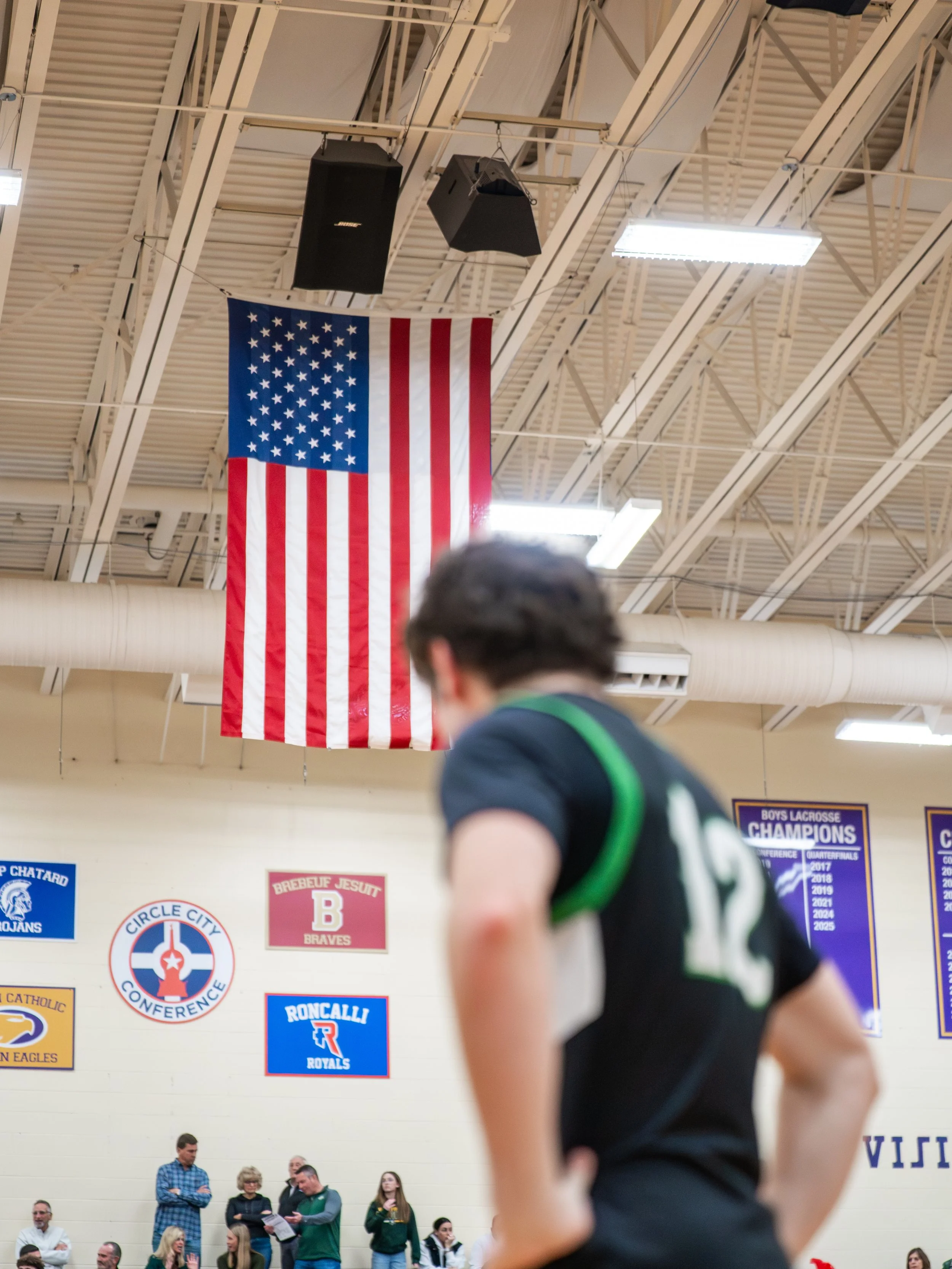 A person with dark hair wearing a black sports jersey with the number 12 stands with hands on hips in a gymnasium. American flags and various school banners are displayed on the wall in the background, along with a group of people.