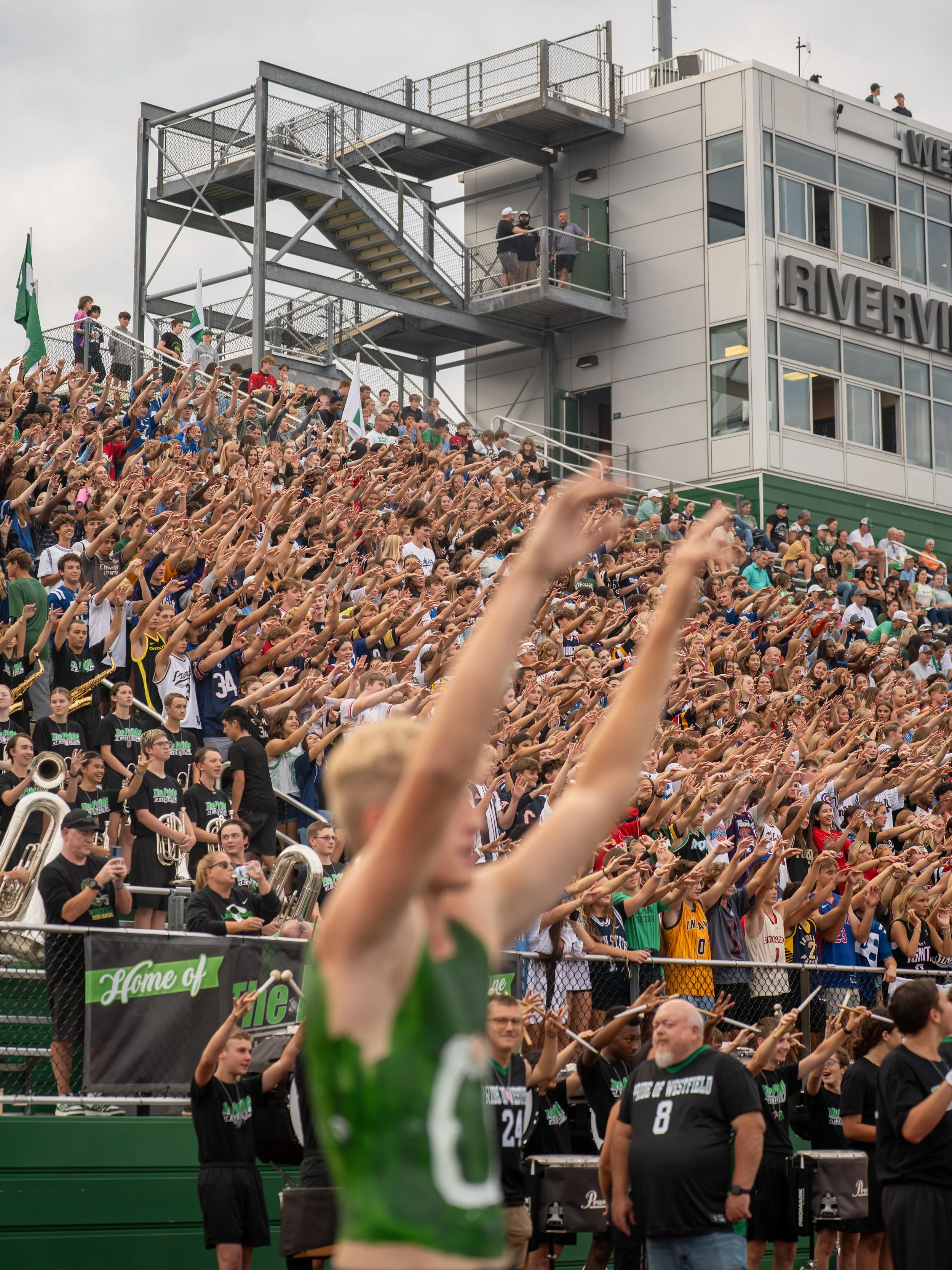 A large crowd of people at a sports stadium, raising their arms in cheer, with some holding flags. A woman in a green sports jersey is in the foreground, slightly blurred, with others behind her participating in the cheer. The stadium has a multi-lev