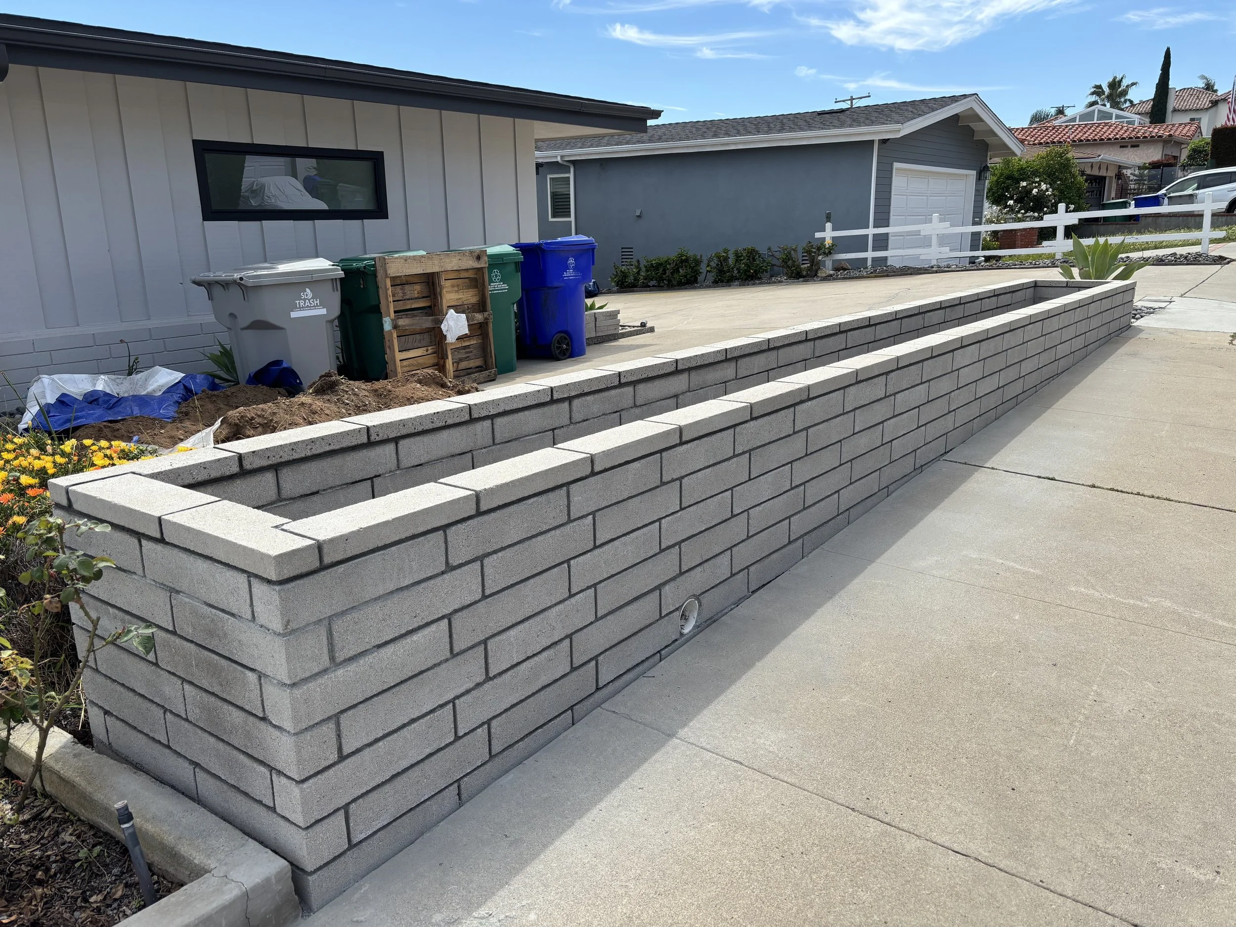 A newly constructed gray brick retaining wall along a sidewalk in a residential area with houses, plants, and trash bins visible in the background.