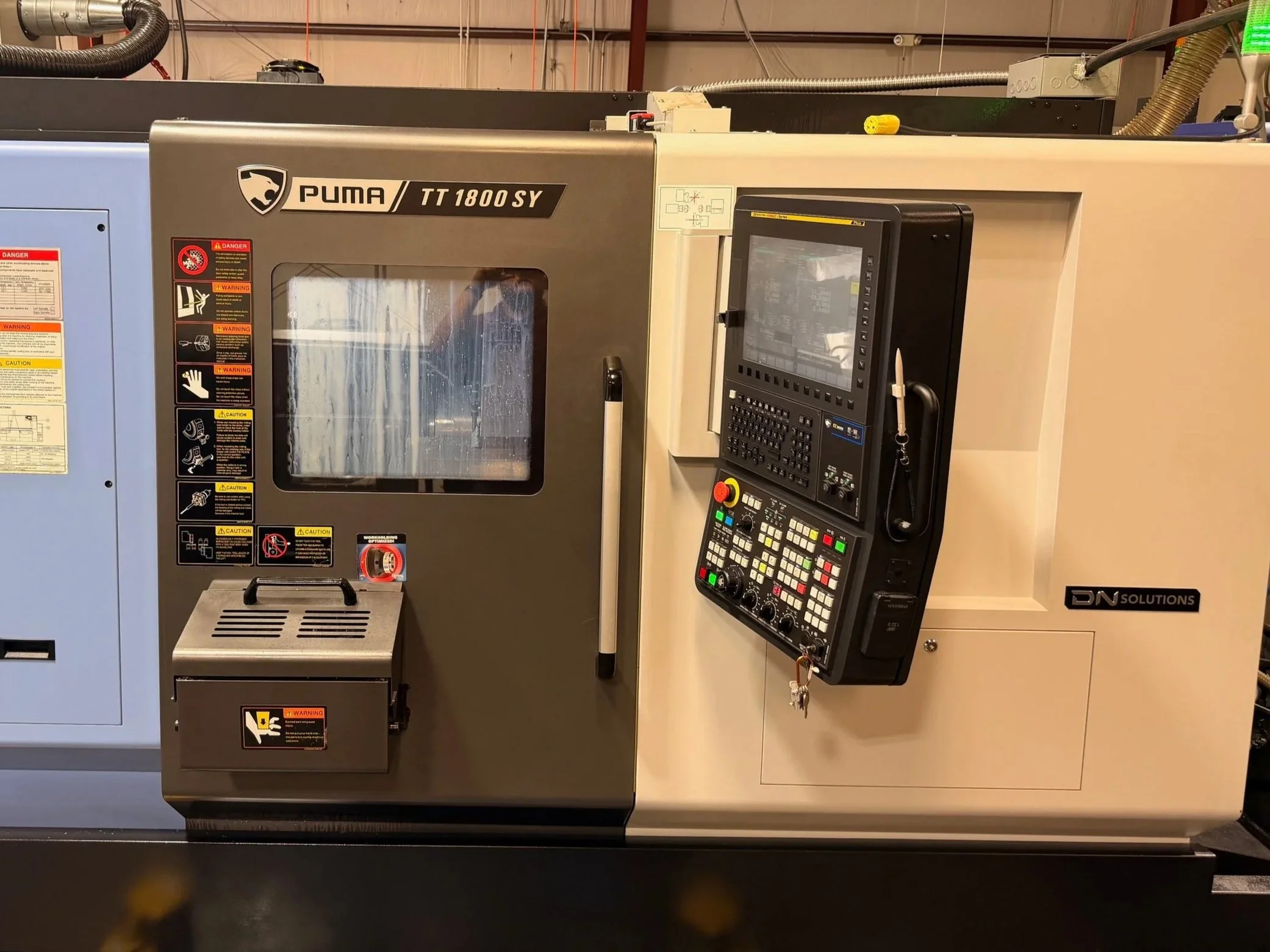 Computer numerical control (CNC) machine used for manufacturing, labeled 'PUMA TT 1800SY,' with control panel, safety warnings, and a window viewing the interior.