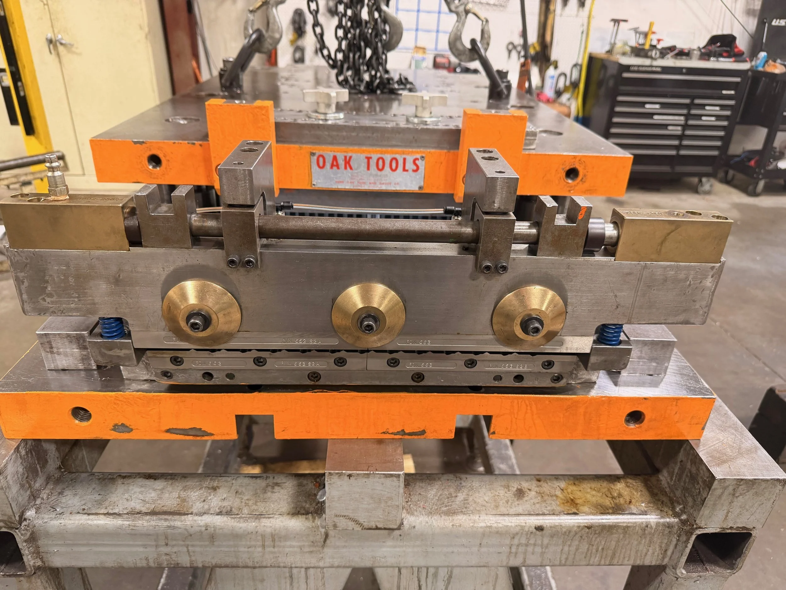 A metal mold fixture with orange blocks labeled 'OAK TOOLS', brass wheels, and various screws and components, set on a worktable in a workshop.
