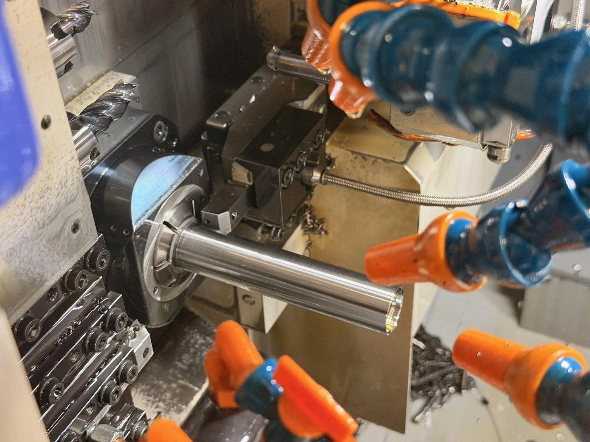 Close-up of a metal lathe machine with a long cylindrical metal piece being machined, various clamps and mechanical parts, and coolant nozzles with orange and blue nozzles.