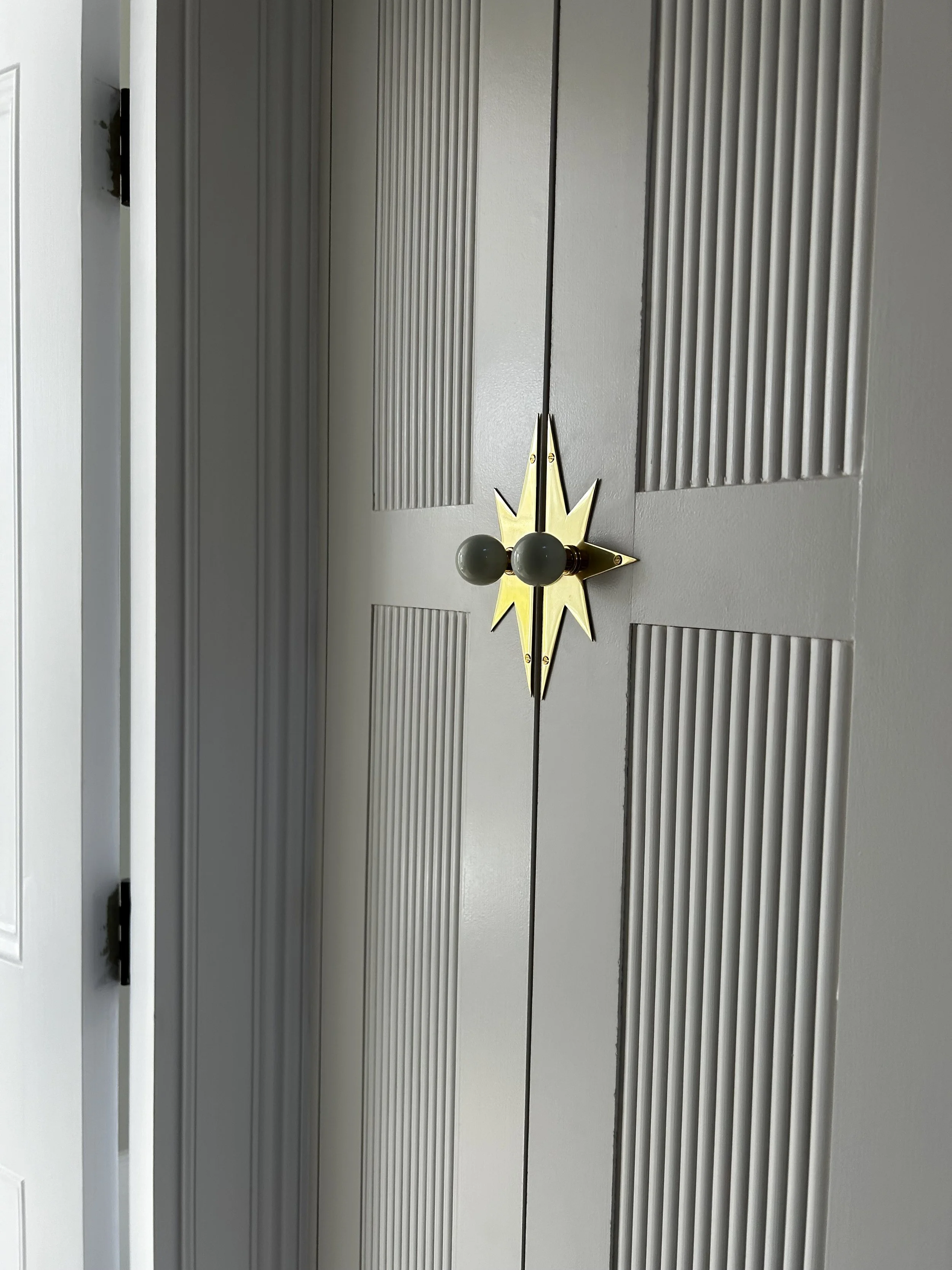 Interior view of a sliding closet door with a vintage starburst-style brass and gray door handle. The door has vertical grooves and is painted light gray.
