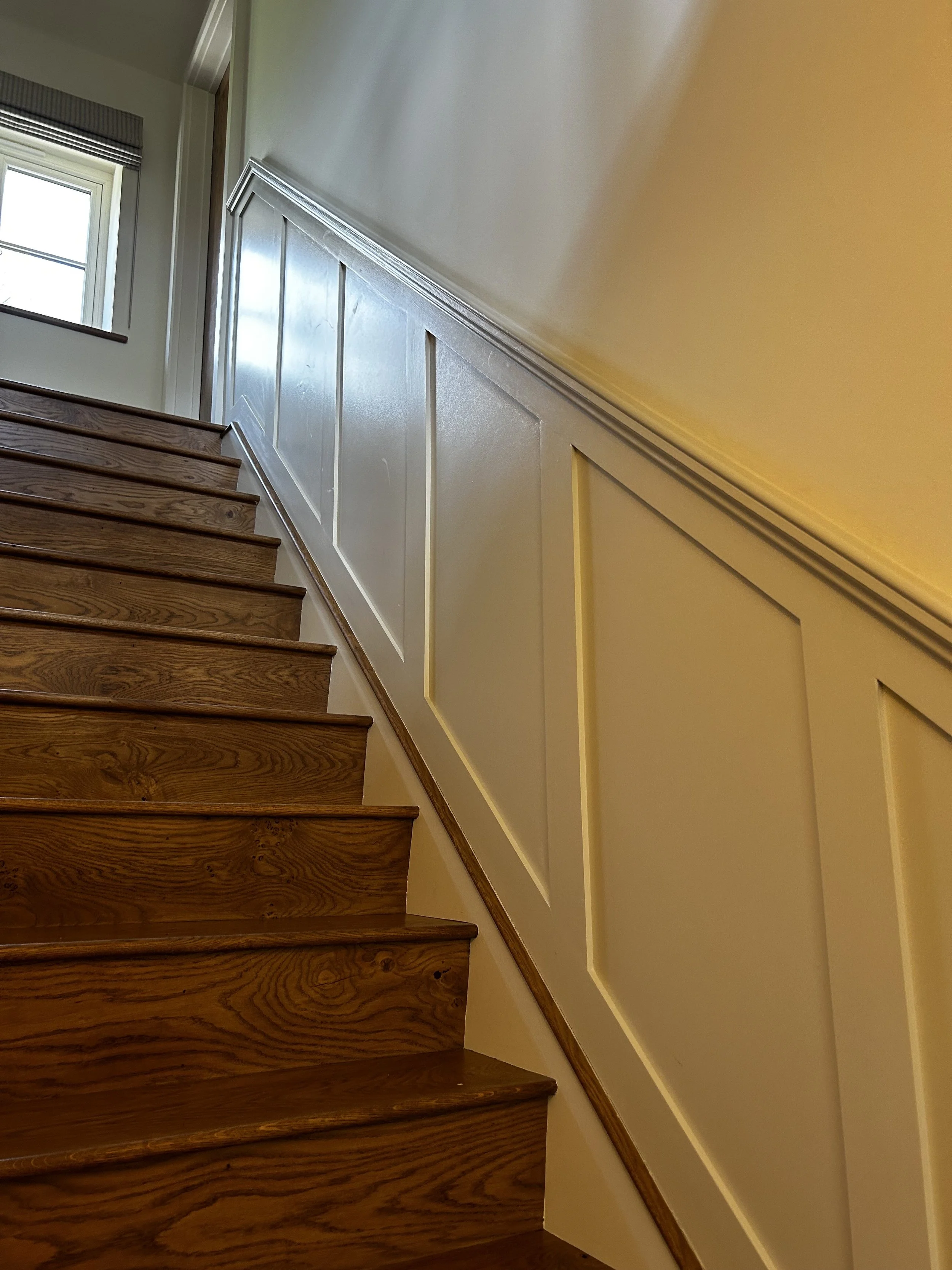 Wooden stairs with white paneled wall on one side and a window at the top of the stairs.
