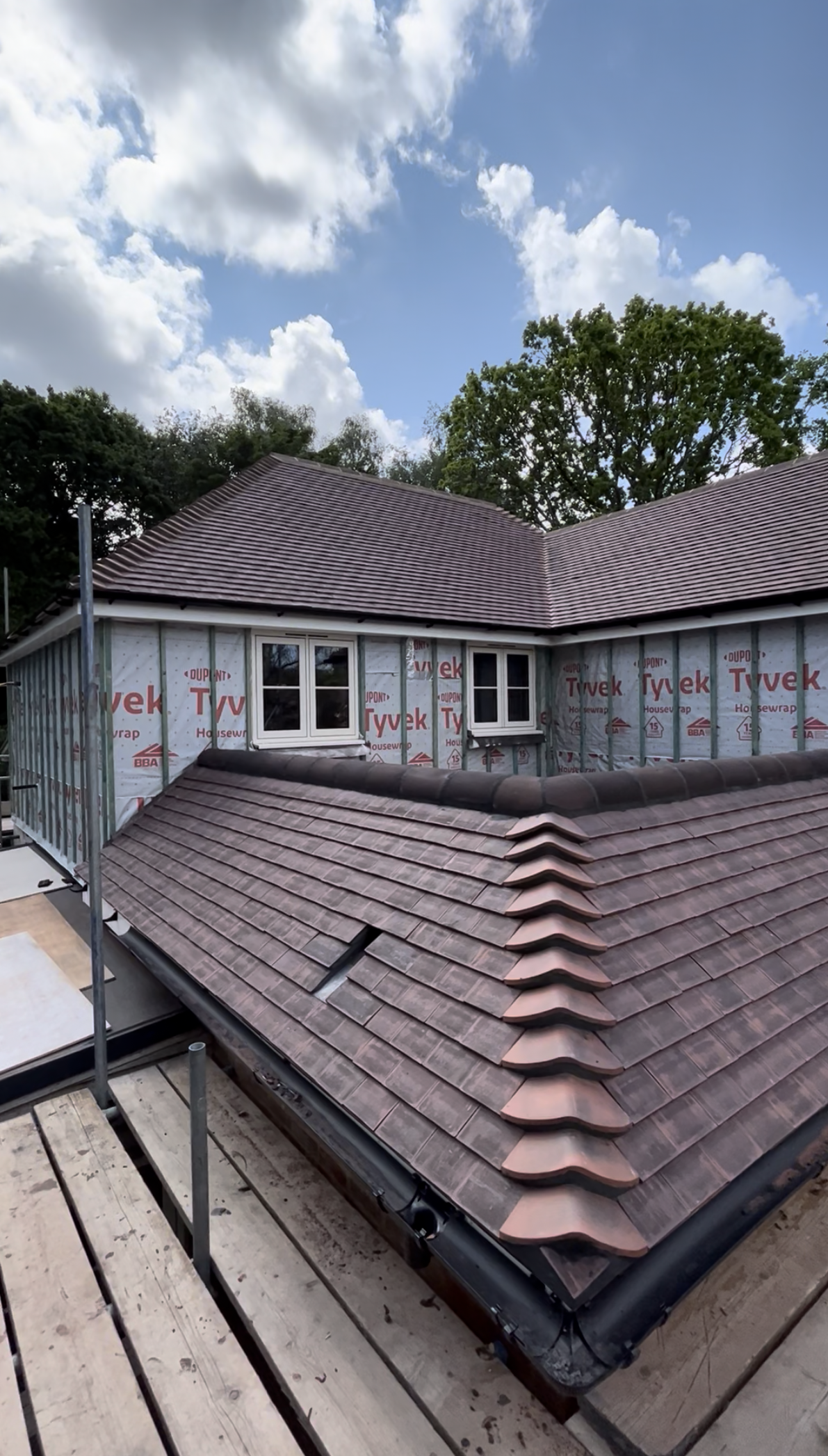 Residential roofing under construction with new roof tiles installed on a sloped roof, windows on a partly covered house, and blue sky with clouds in the background.