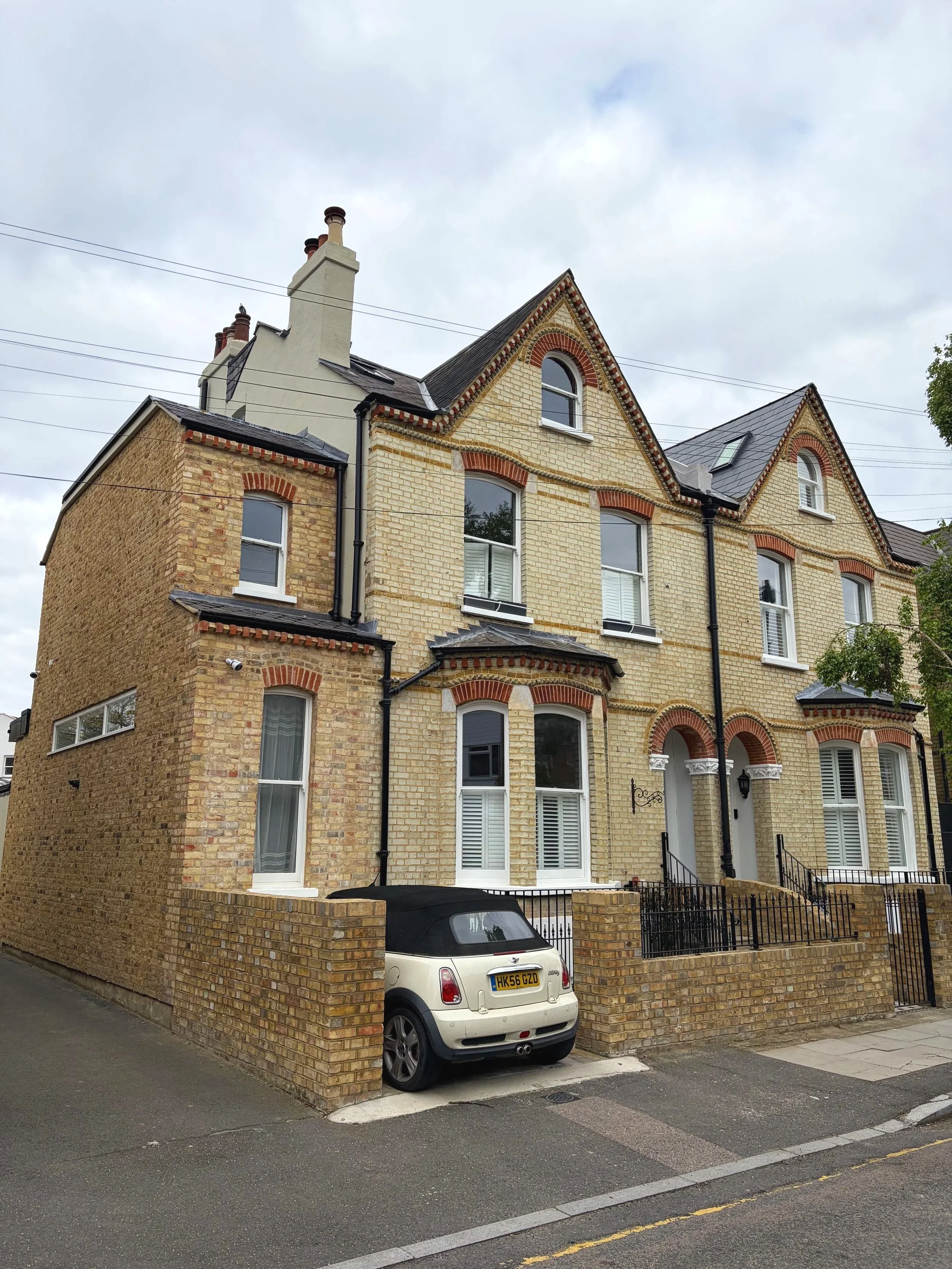 A multi-story brick house with arched windows and a small white car parked in front surrounded by a brick wall.