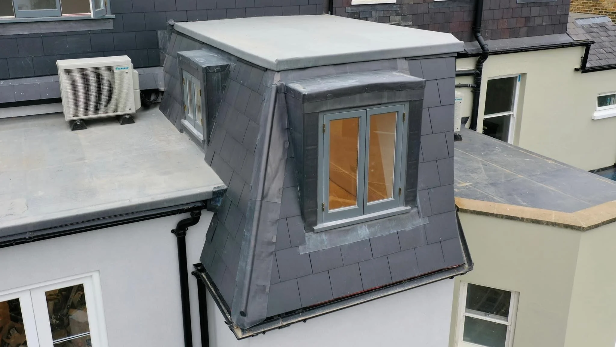 Close-up aerial view of a house rooftop with a dormer window, an air conditioning unit, and surrounding parts of the roof and neighboring roof structures.