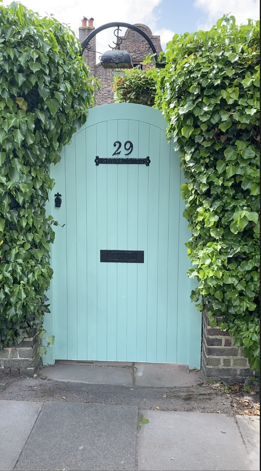 A light turquoise wooden door with the number 29 on it, surrounded by lush green ivy, with a black mailbox and a bell on the door, and a historical brick building in the background.
