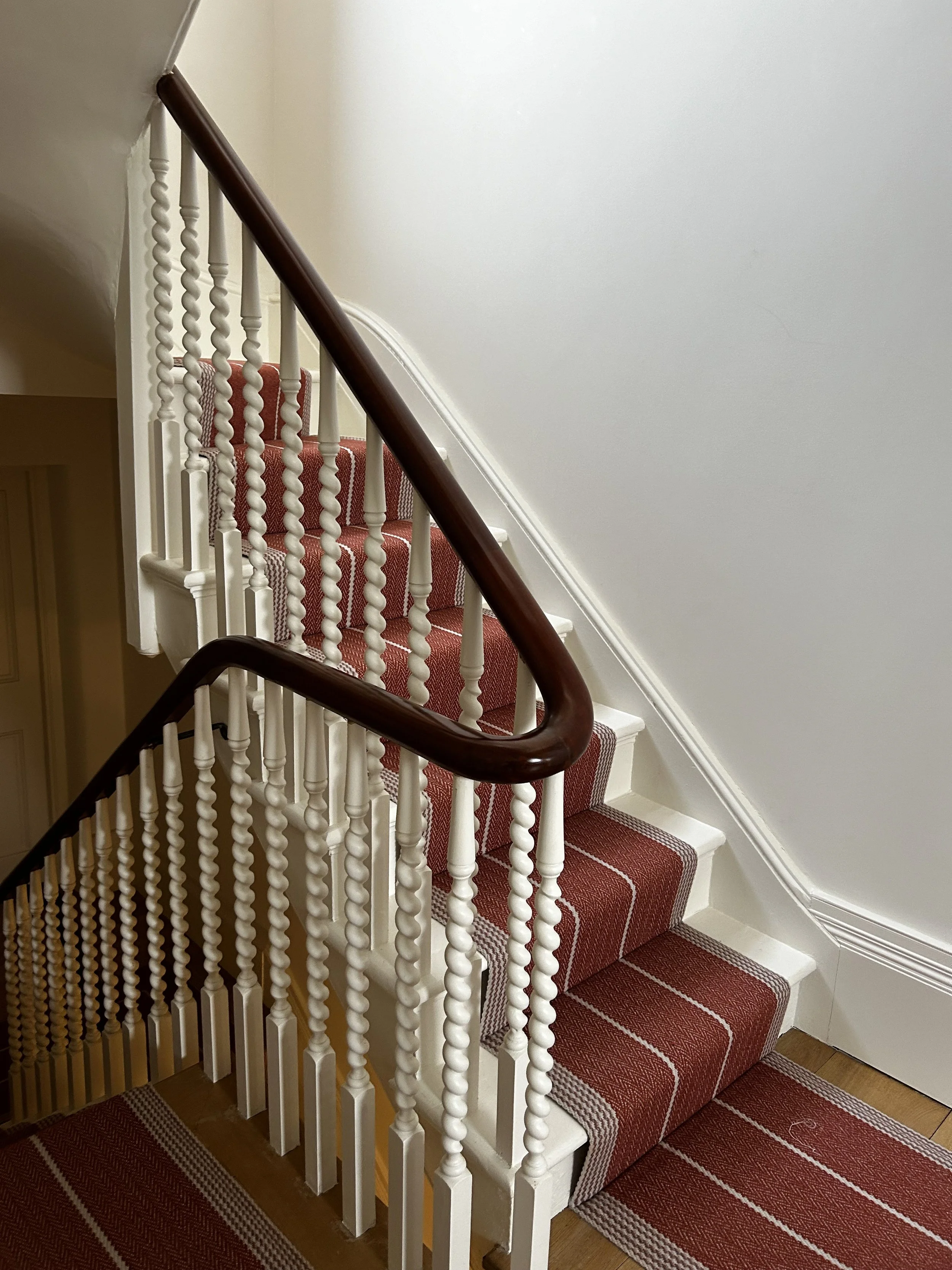 Interior view of a staircase with a red and white striped carpet, white wooden balusters, and a dark brown wooden handrail.