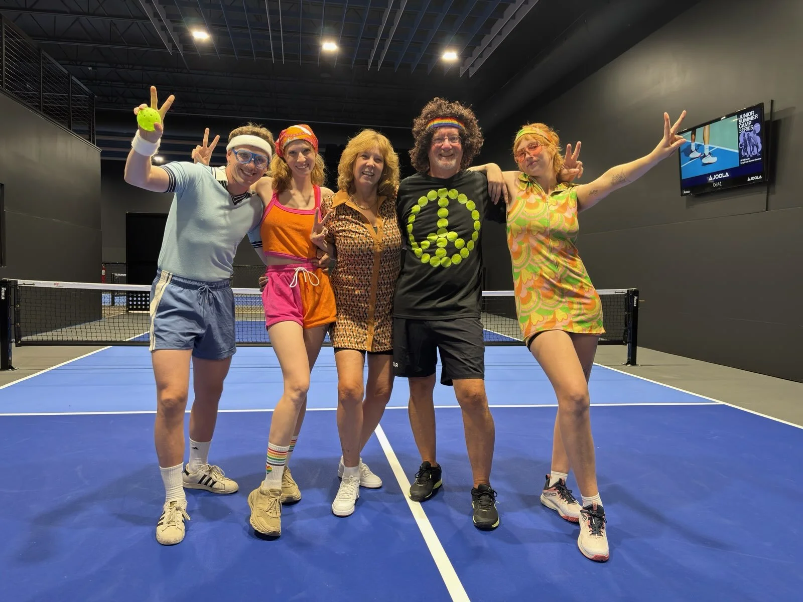 Five people dressed in colorful 70s inspired tennis outfits posing on a blue indoor tennis court, making peace signs and smiling @the Picklr Bozeman.