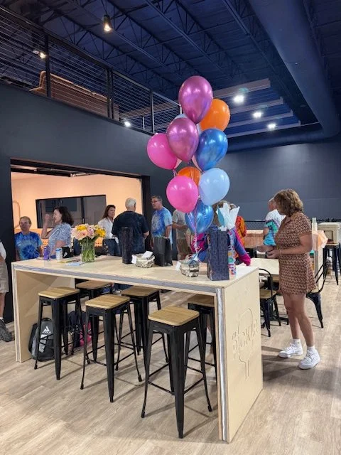 Party scene with balloons, flowers, and several people gathered around a white counter in a modern indoor space.