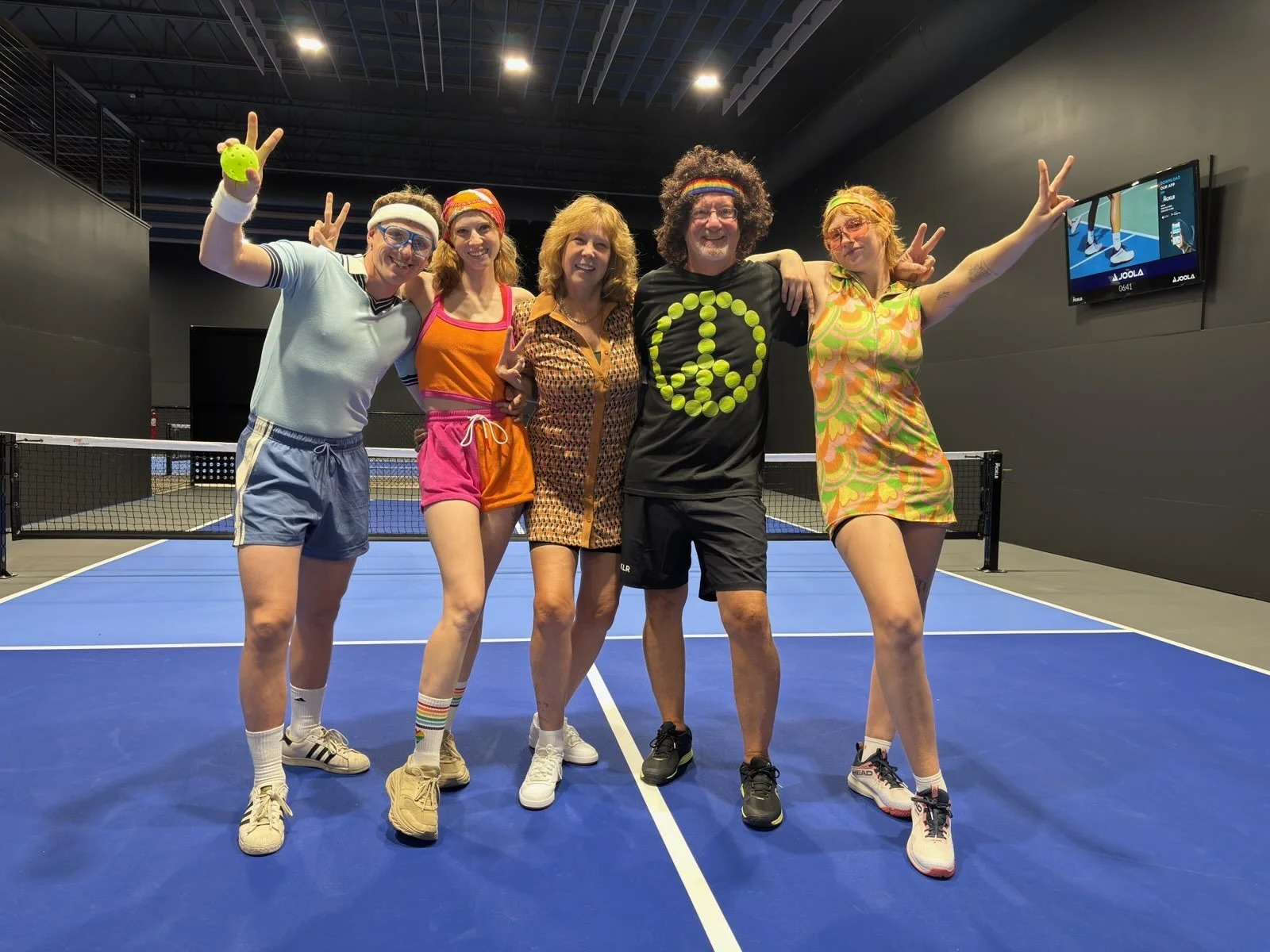 Five people dressed in colorful 70s inspired tennis outfits posing on a blue indoor tennis court, making peace signs and smiling @the Picklr Bozeman.