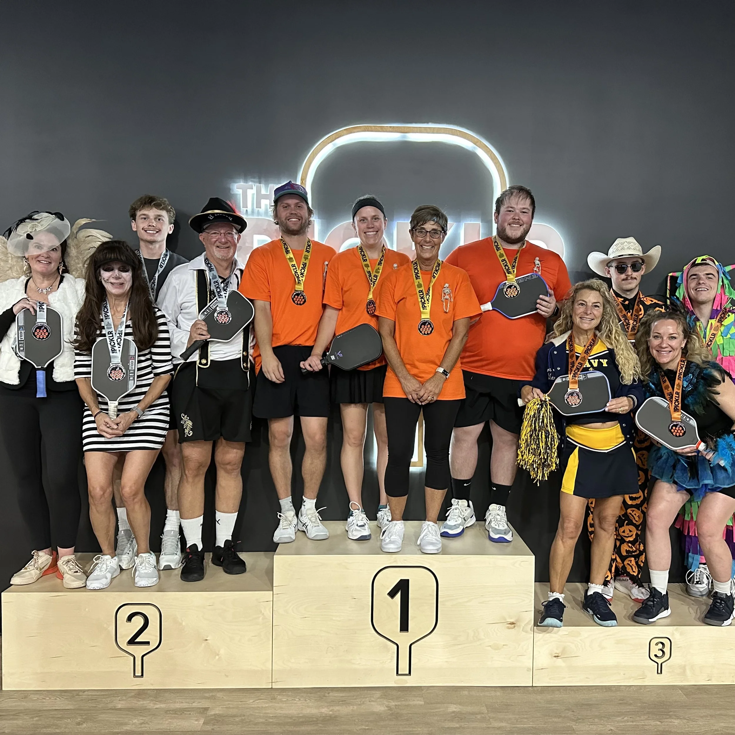 Group of people dressed in costumes and athletic attire standing on winners' podium with medals around their necks, celebrating a victory at a Bozeman Pickleball event at the Picklr