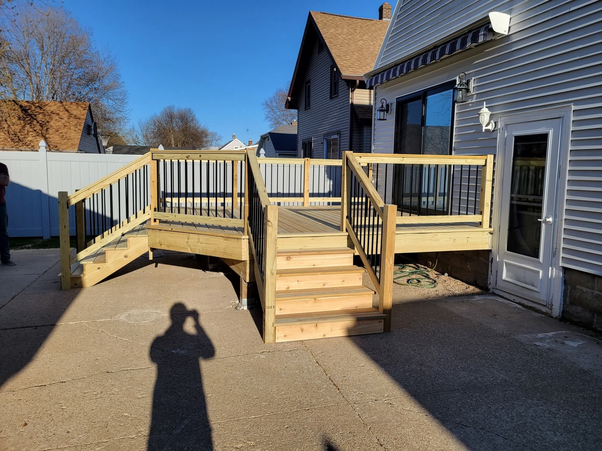 New wooden deck with black metal railings attached to the back of a house, with stairs leading down to a concrete patio, in a residential neighborhood.