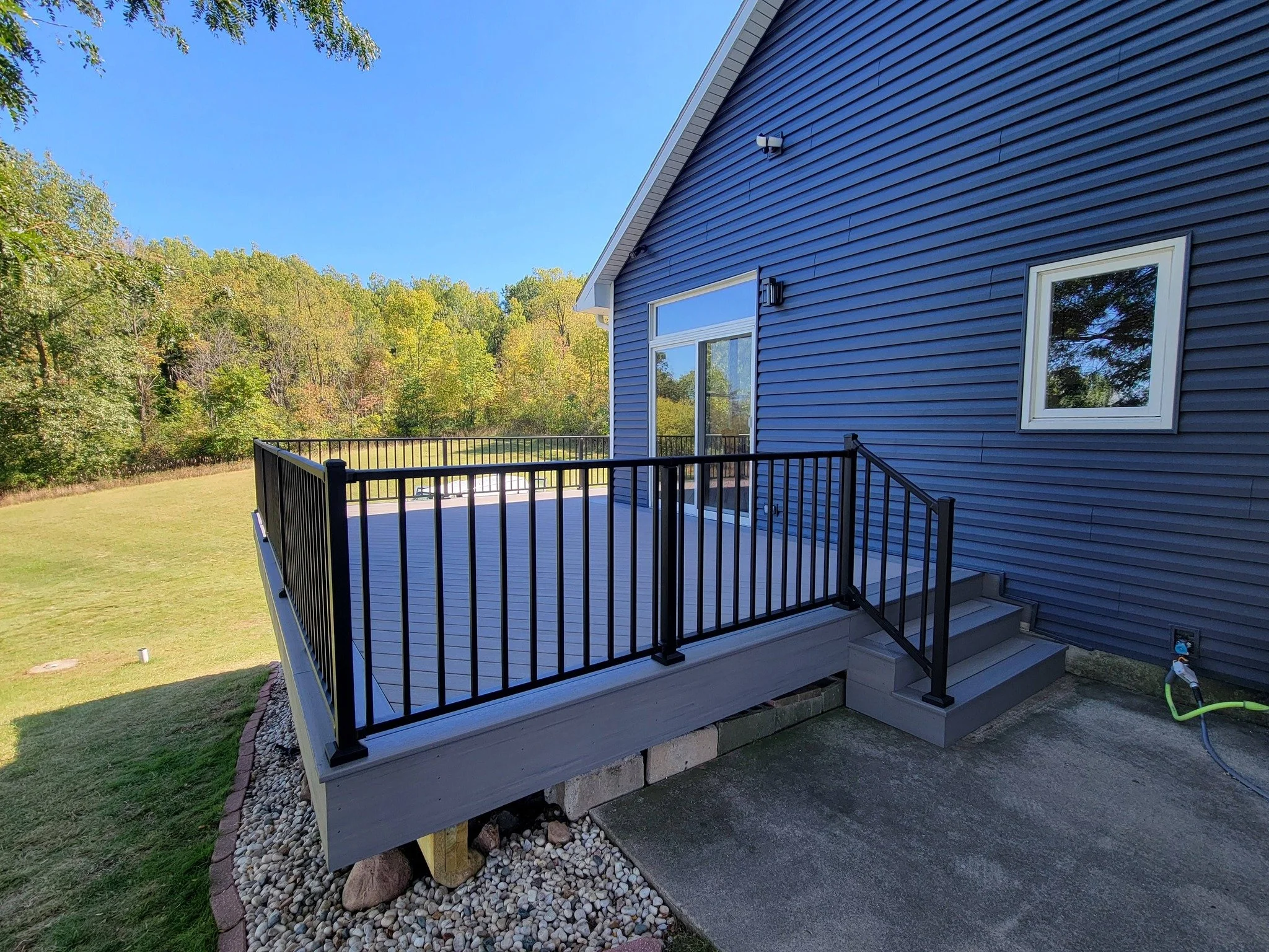 New light gray composite deck with black aluminum railing attached to a blue house with white trim, stairs leading down, outdoor faucet with hose, and a grassy backyard with trees.