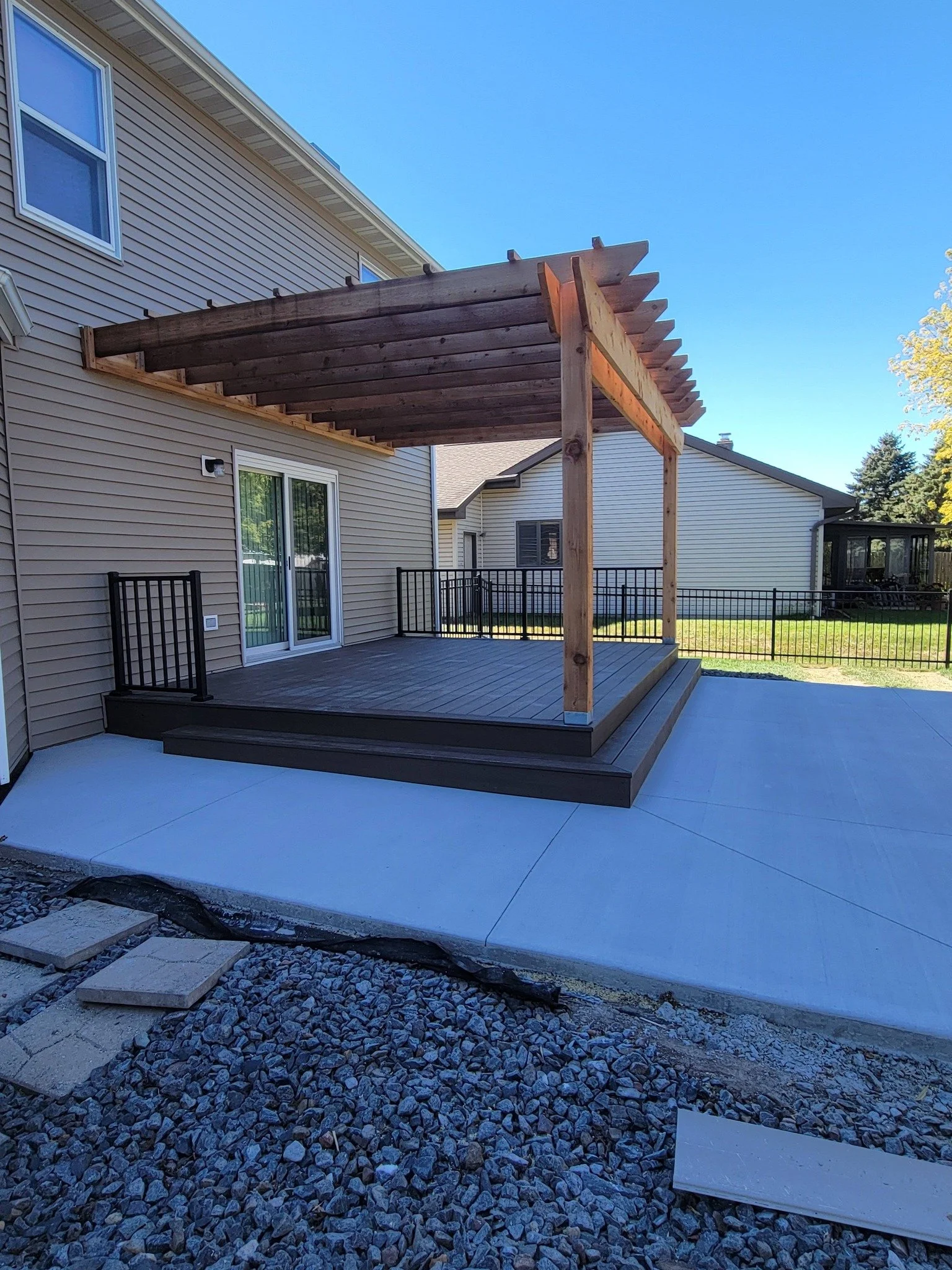Backyard patio with a new wooden pergola, a concrete slab, and a raised deck with black aluminum railing, adjacent to a beige siding house with sliding glass door.