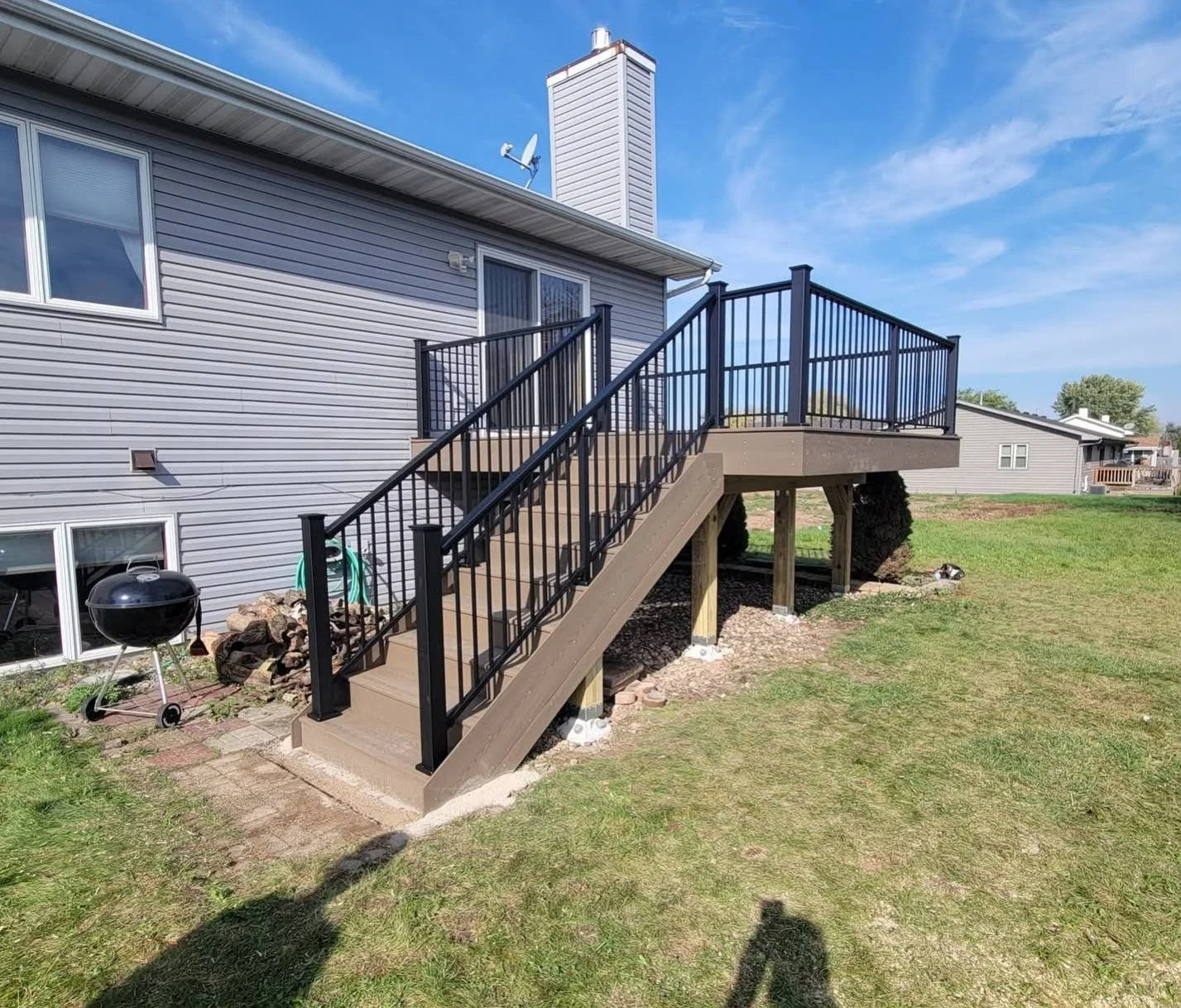 Newly built elevated wooden deck with black metal railings attached to gray house with sliding glass door, located in a backyard with green grass and neighboring houses in the distance.