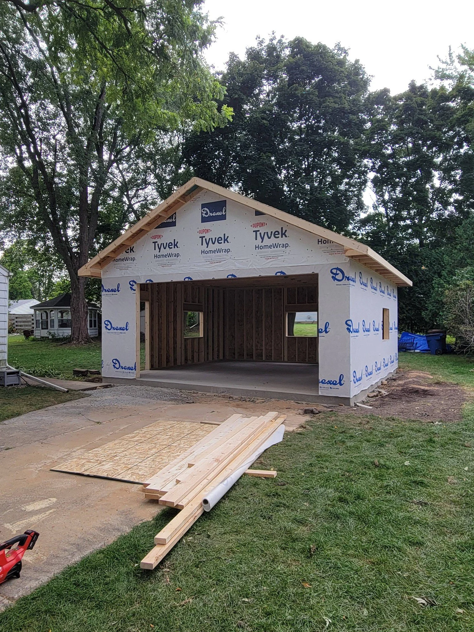 A new constructed garage before the finishing touches. The structure is in a grassy yard, with building materials and tools on the ground nearby. Trees and other houses are visible in the background.