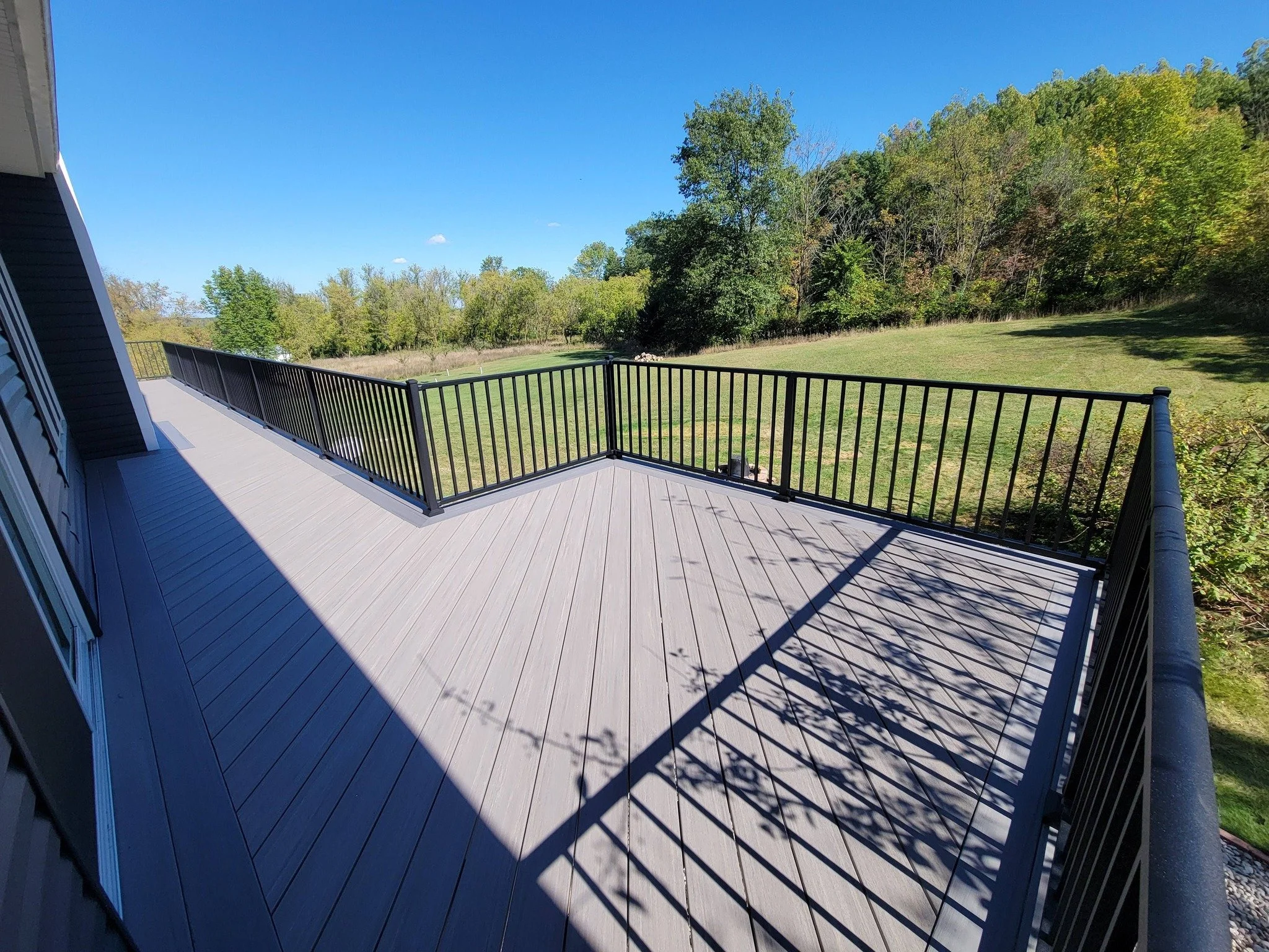 A spacious outdoor deck with light gray composite flooring and black aluminum railing, overlooking a grassy yard and tree-lined landscape on a sunny day.
