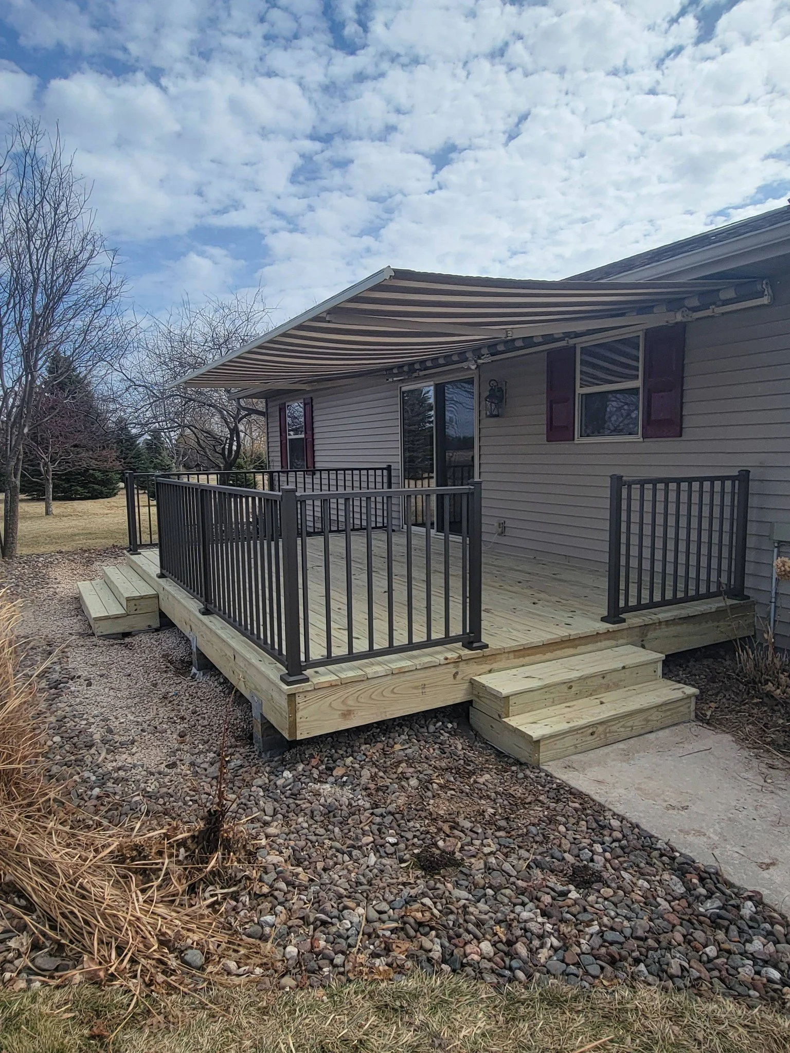 New wooden deck with black aluminum railing attached to the back of a house, with steps leading down to a gravel yard and an overcast sky.