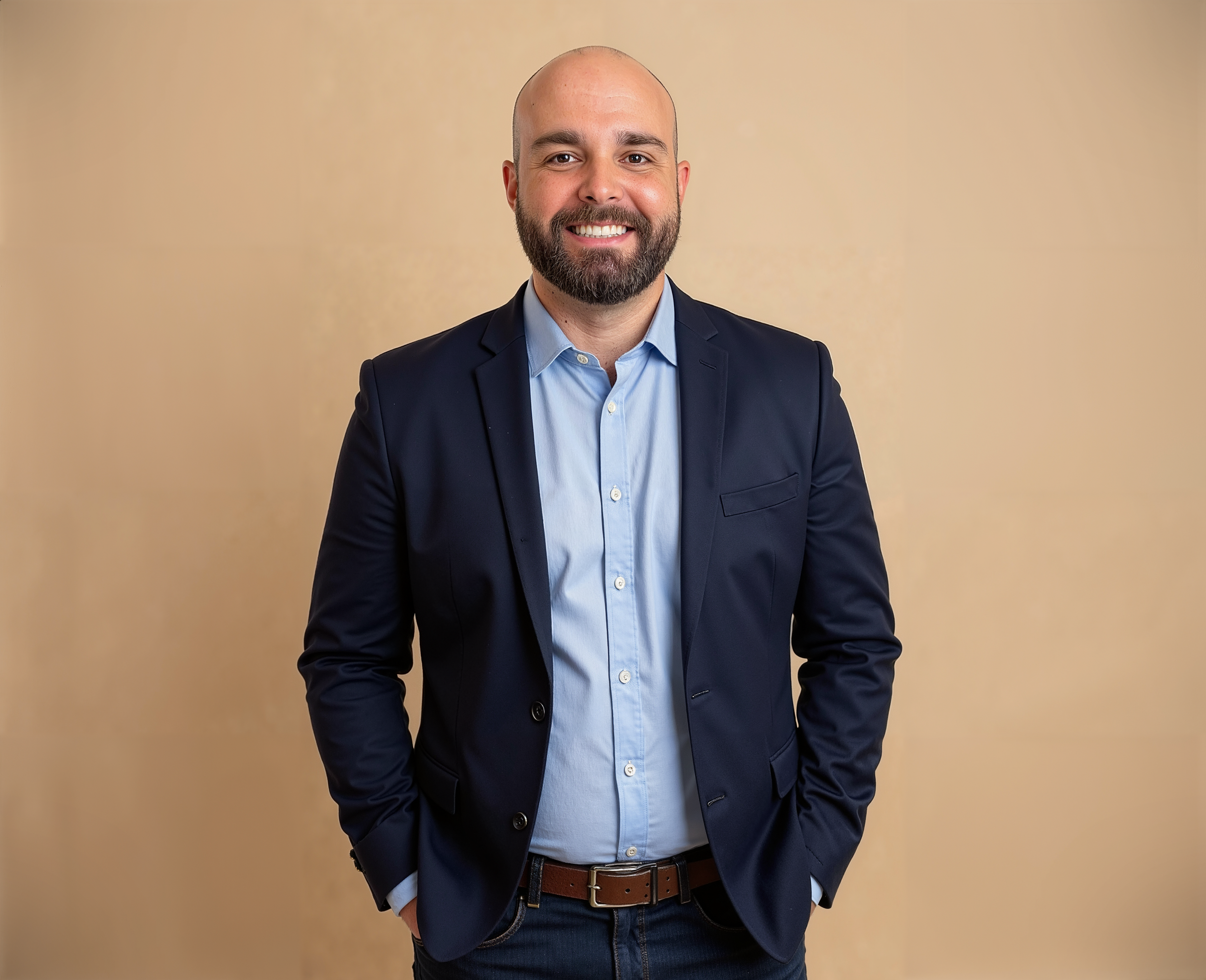A smiling man with a beard dressed in a navy suit jacket, light blue shirt, and brown belt, standing against a plain beige background.