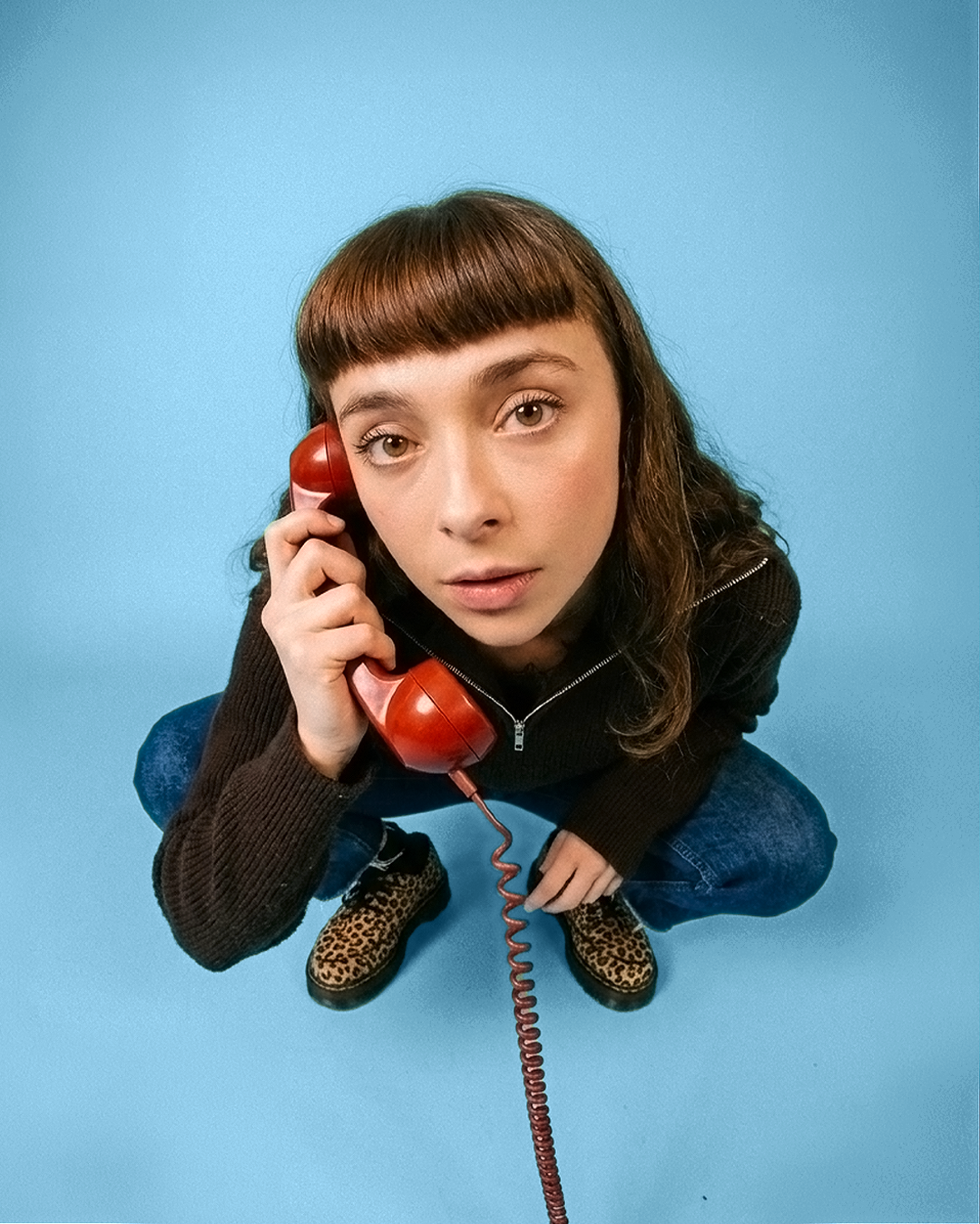 Une photo de moi, des cheveux auburn et un maquillage naturel, portant un pull marron et des chaussures à imprimé léopard, tenant un téléphone rétro rouge à l'oreille, assise sur un sol bleu.