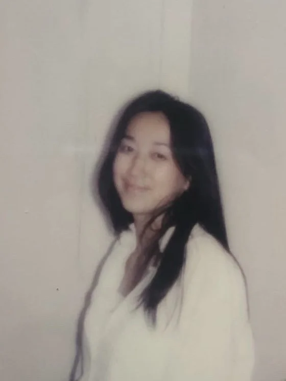 A woman with long black hair, smiling in front of a plain wall, wearing a light-colored top.