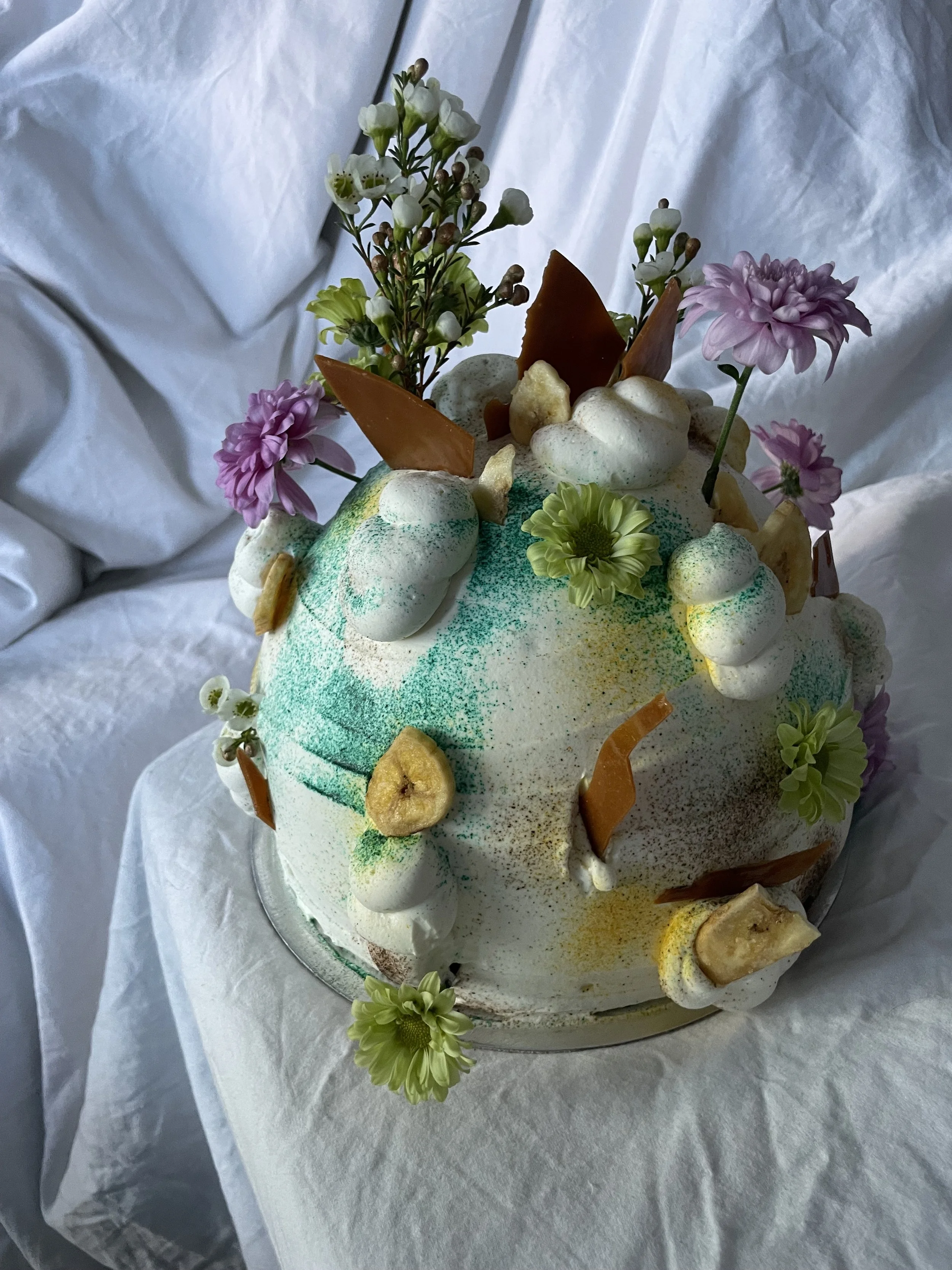 A decorated cake with white frosting, colorful sprinkles, edible flowers, caramel shards, and banana slices, set on a white cloth.