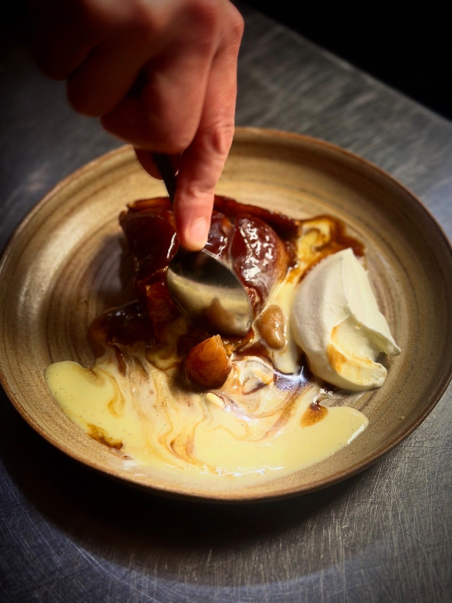 Hand adding spoonful of caramel sauce to a bowl containing whipped cream, nuts, and caramelized fruit.