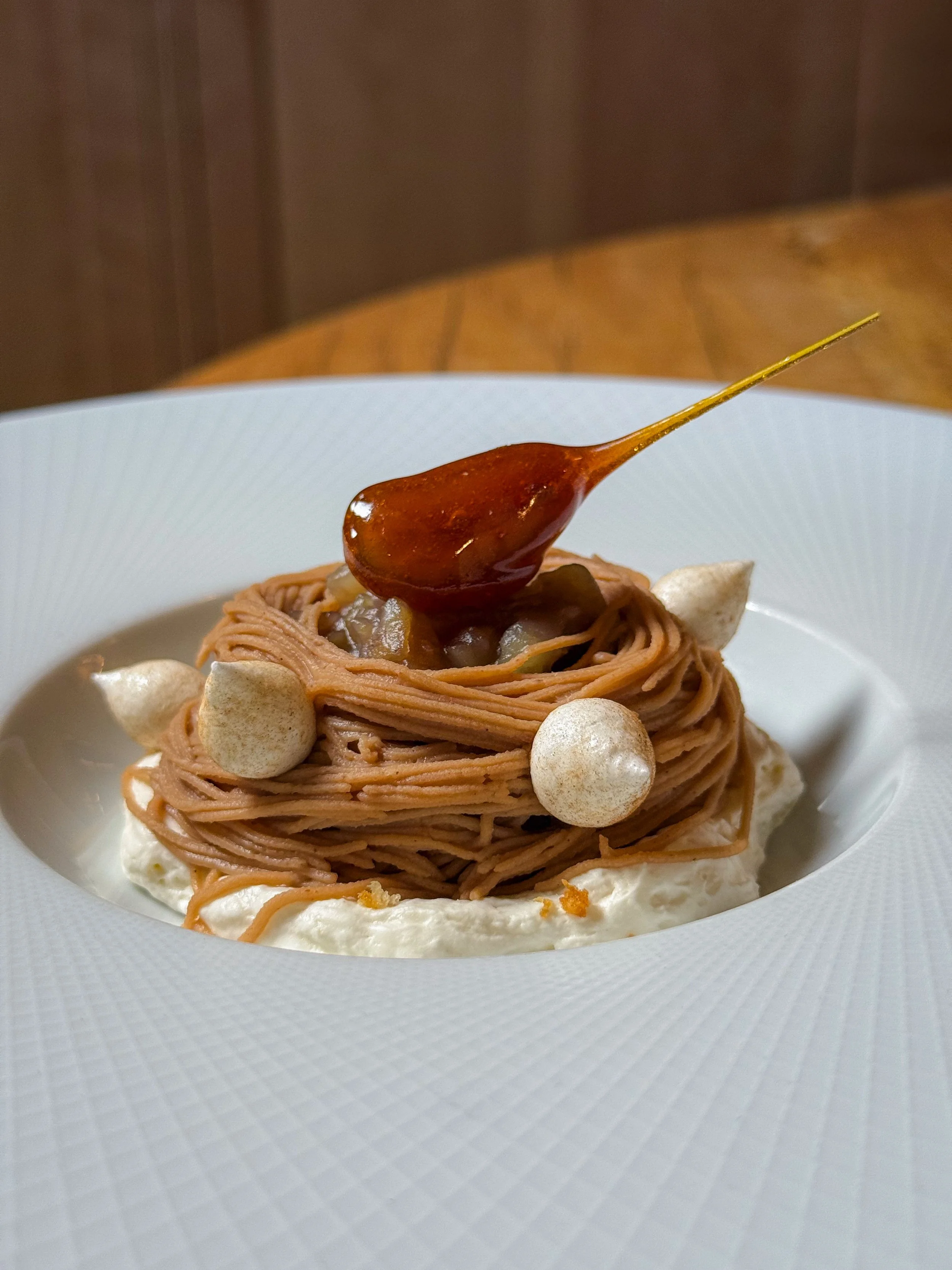 A plated gourmet dessert featuring a nest of chocolate spaghetti topped with a spoonful of sauce, accompanied by meringue kisses and creamy dollops on a white textured plate.