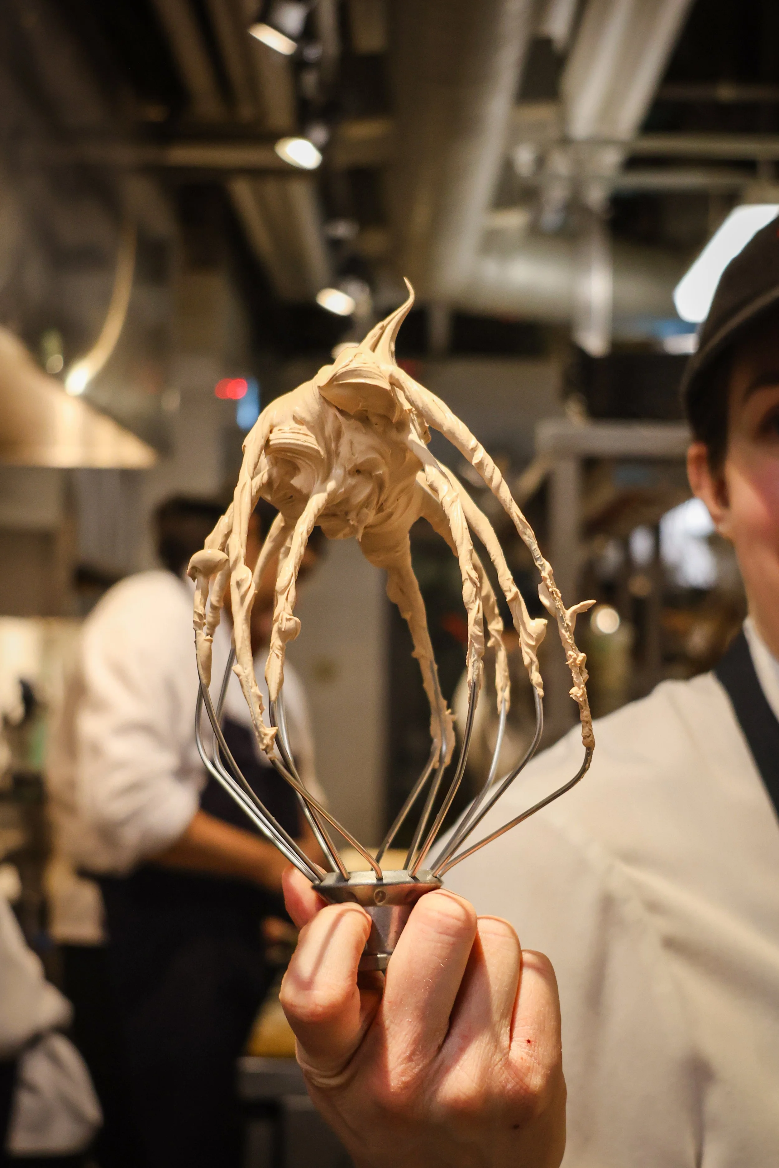 Person holding a metal whisk with a dollop of thick, beige frosting or batter.