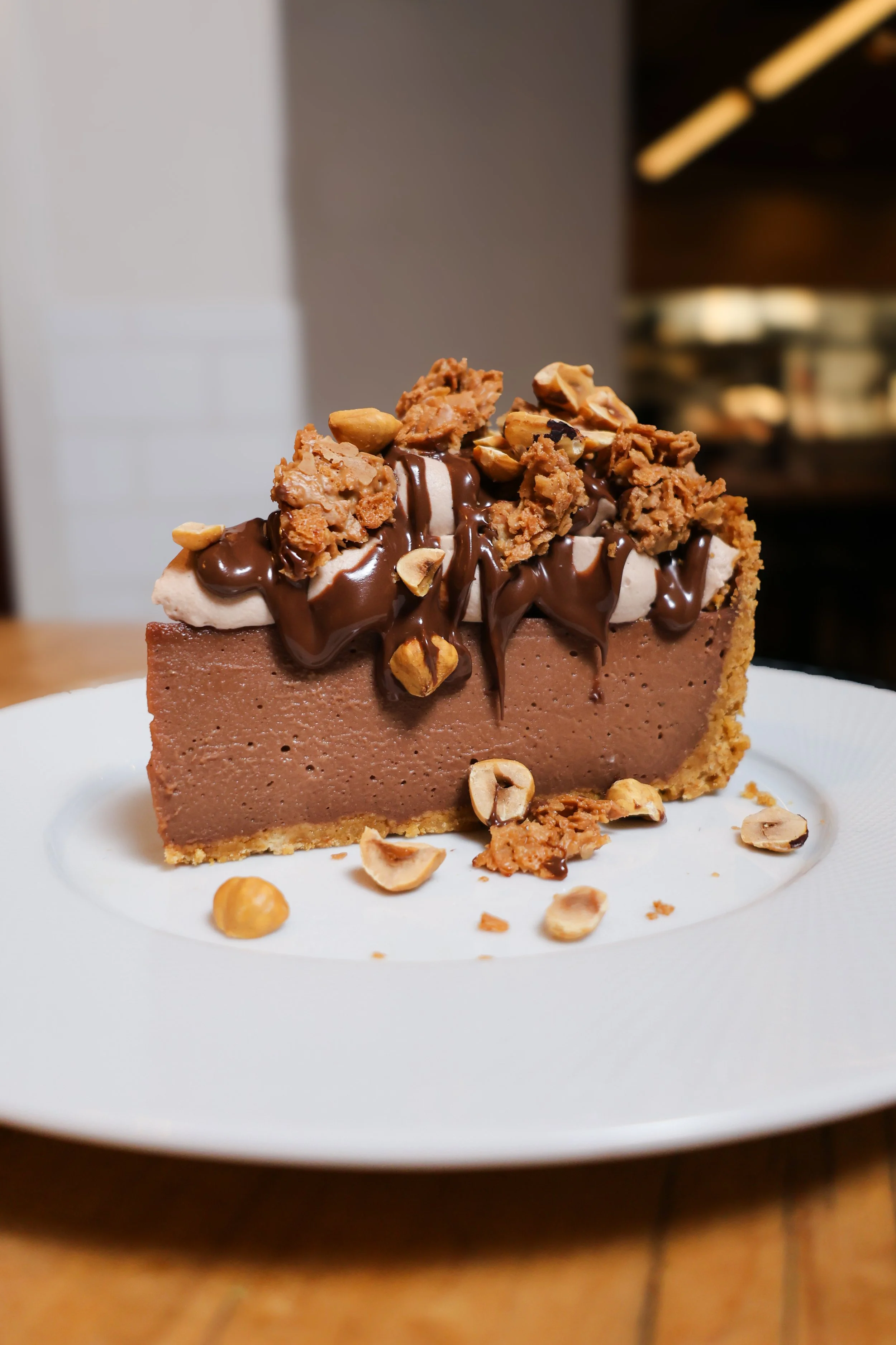 Slice of chocolate cheesecake topped with caramel crumble, chocolate drizzle, and hazelnuts on a white plate.