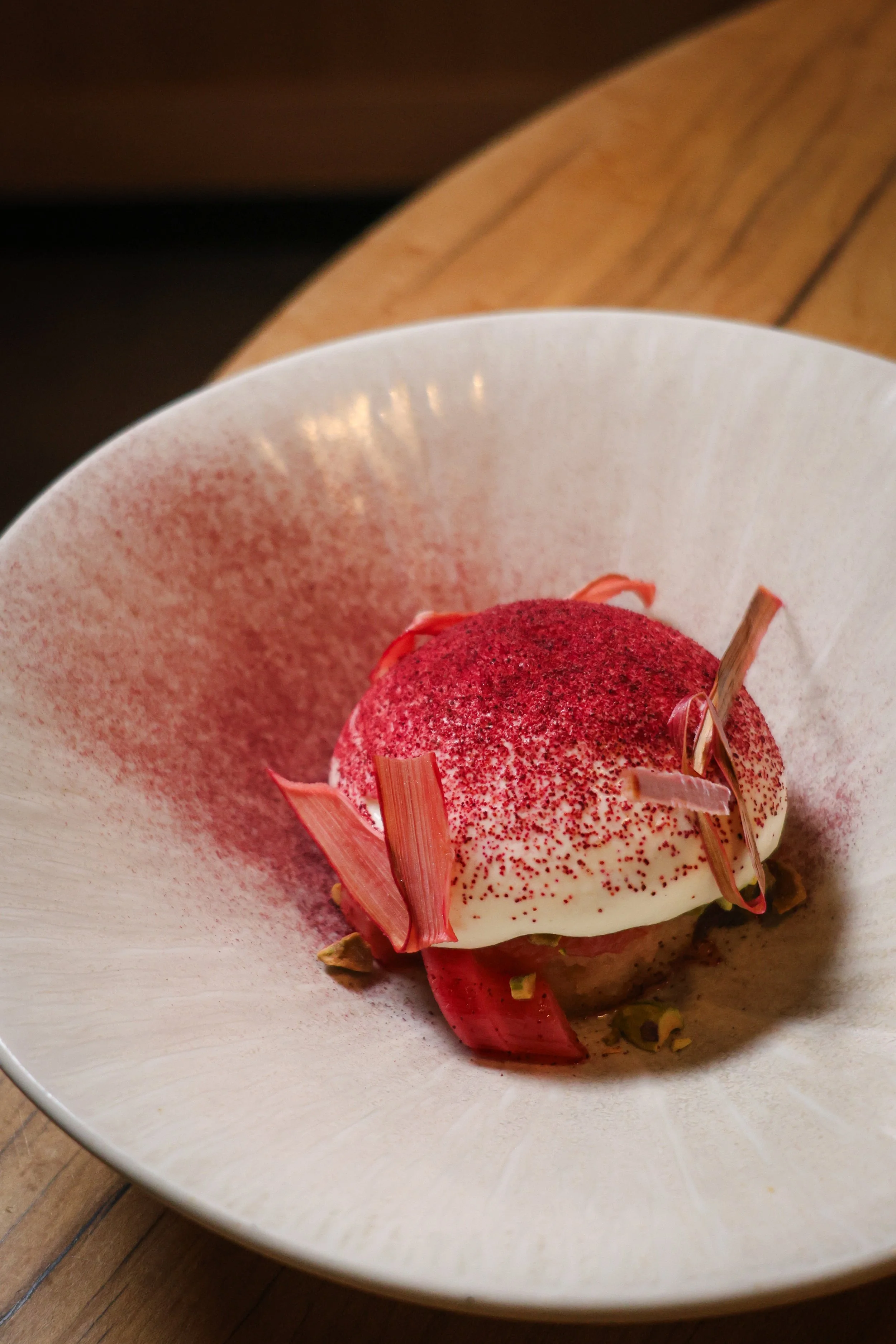 A plated dessert with white creamy base, red powder topping, pinkish-red decorative petals, and chopped nuts in a white bowl on a wooden surface.
