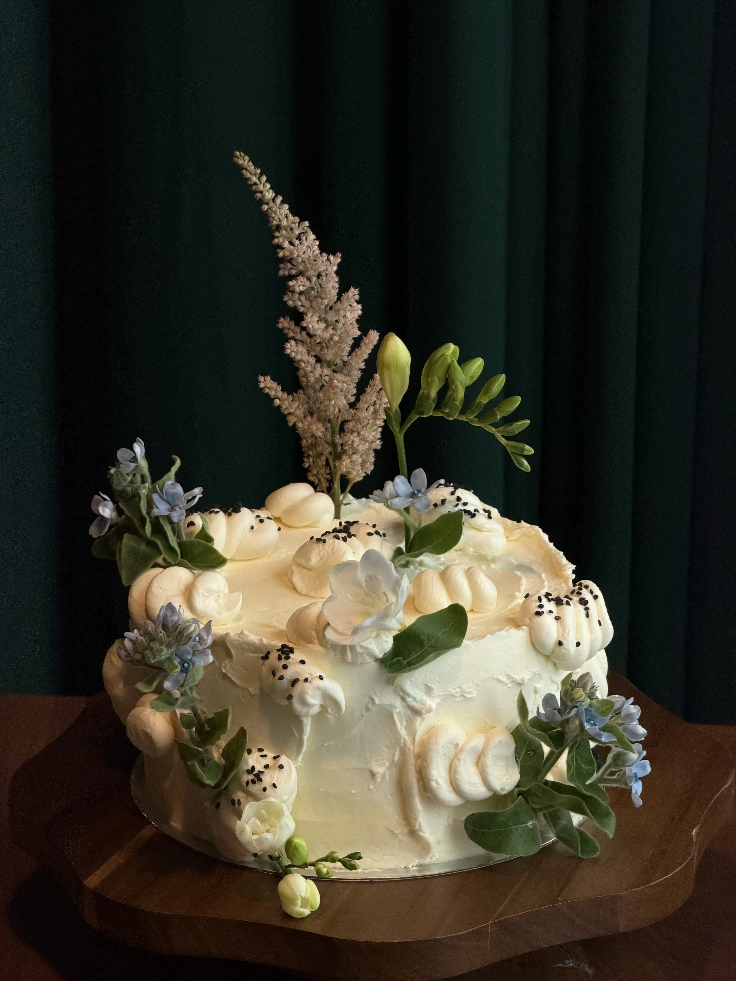 A decorated white cake with flowers and greenery, topped with a tall pinkish flower, small blue flowers, and black sprinkles, placed on a wooden board against a dark curtain background.