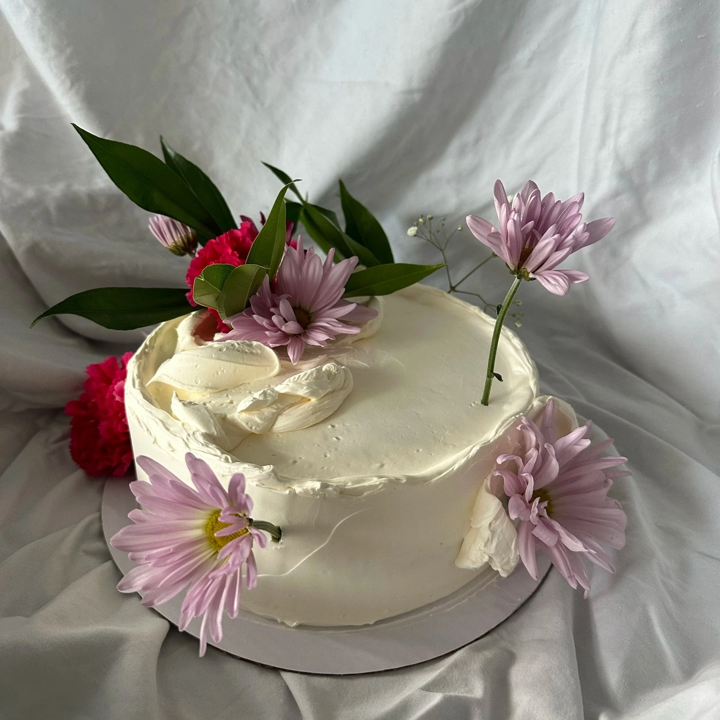 White cake decorated with pink daisies, pink carnations, green leaves, and a small sprig of baby's breath, placed on a white cloth background.