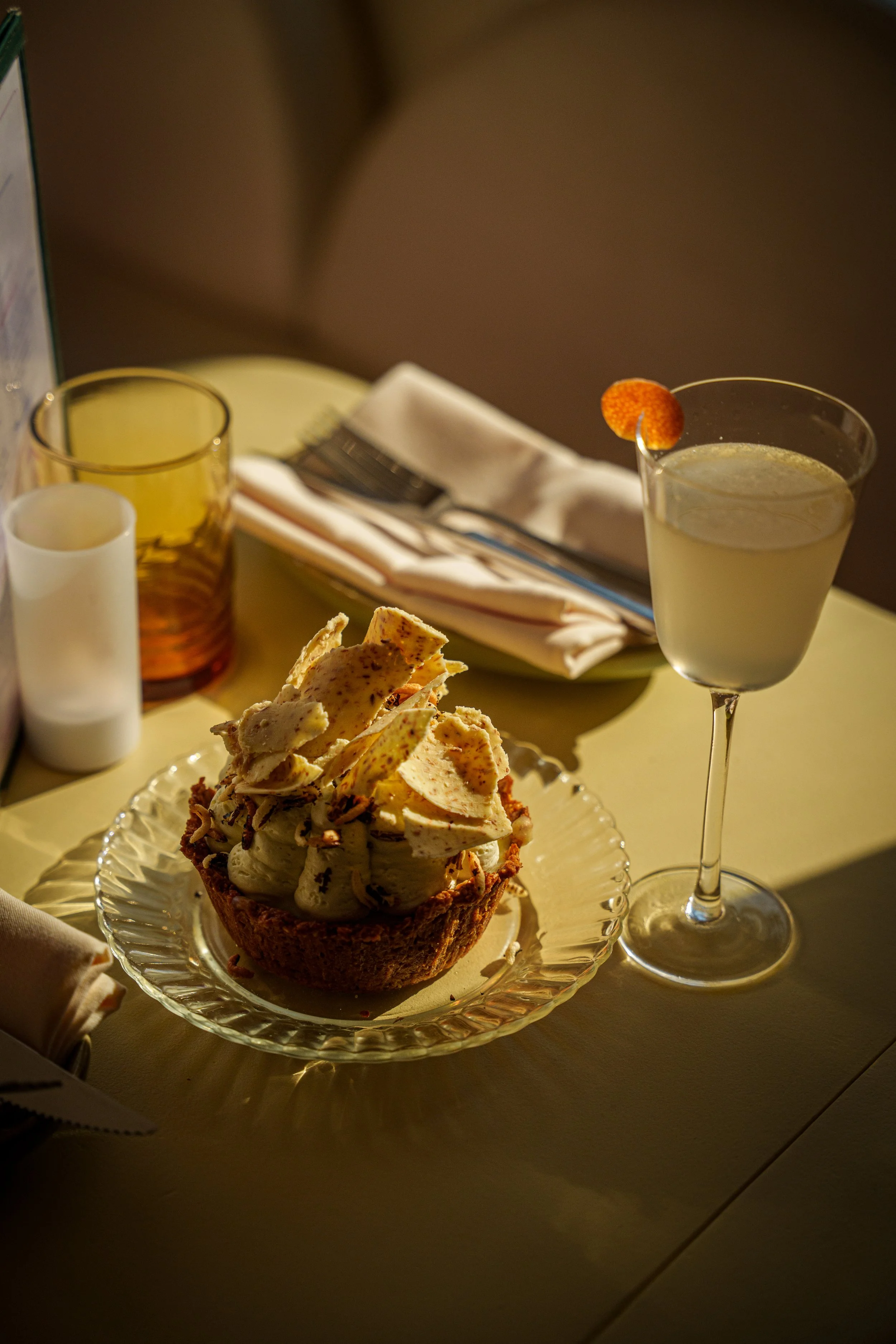 A dessert of ice cream topped with tortilla chips, served on a glass plate, alongside a cocktail with a citrus garnish on a yellow table setting.